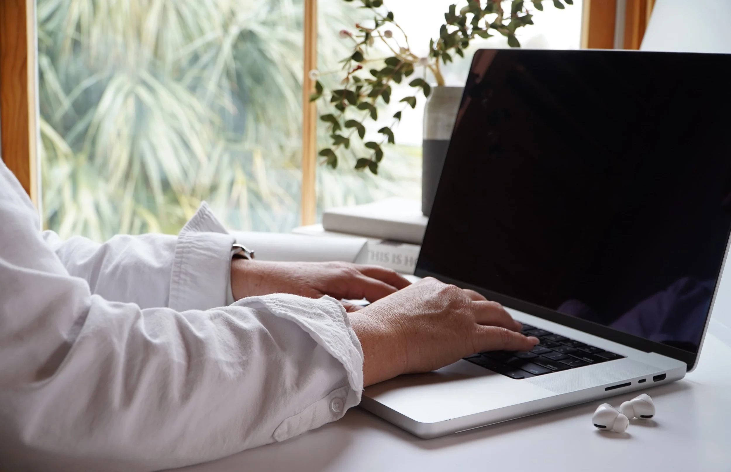 Person typing on a silver laptop with a black screen, sitting at a white desk with wireless earbuds nearby. In the background, a houseplant and books are visible near a window with greenery outside.
