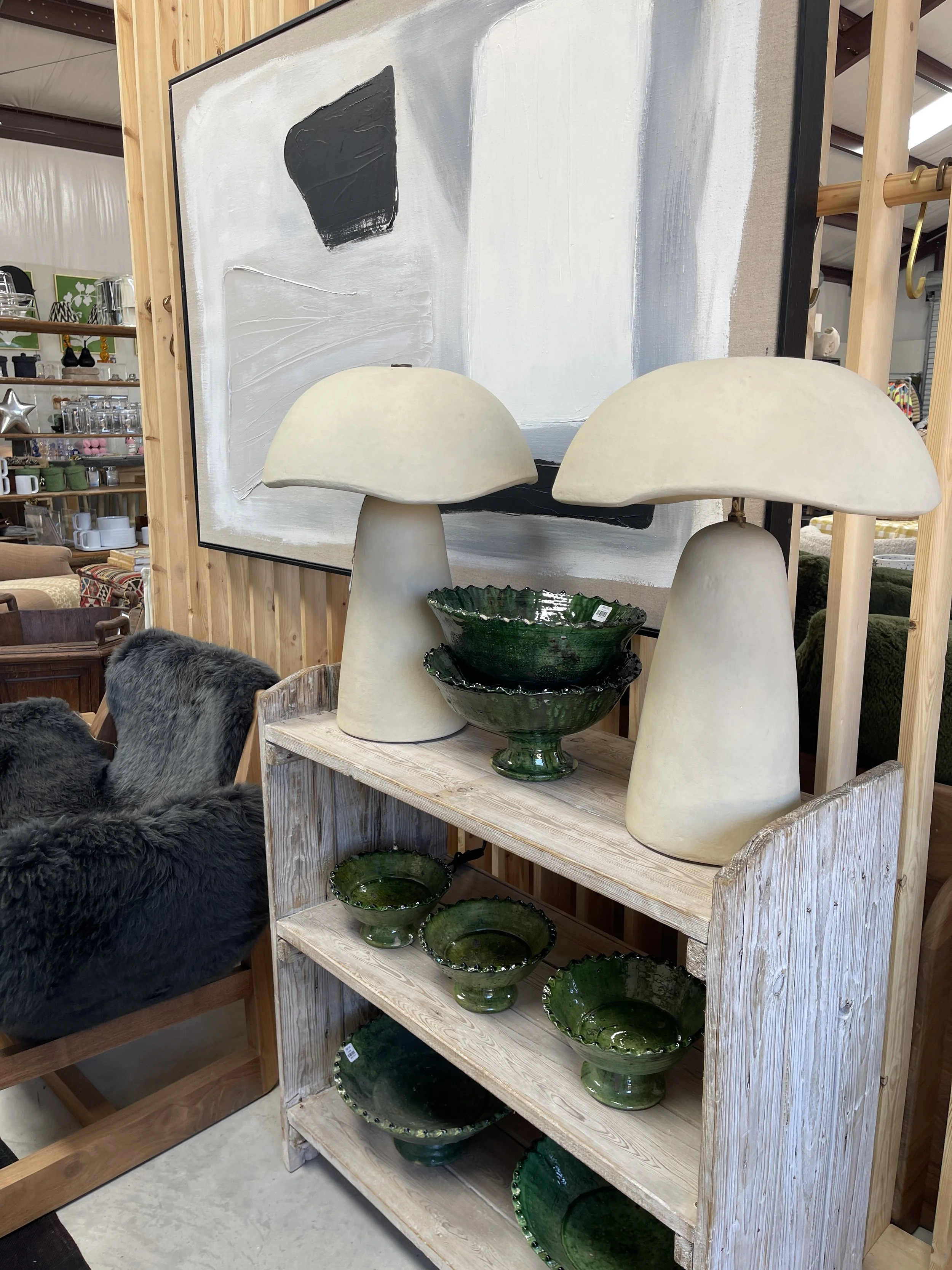 A wooden display shelf holding several green glazed bowls and ceramic lamps resembling mushrooms positioned on top of the shelf. A black and white abstract painting hangs on the wall behind the lamps. The setting appears to be in a home decor or antique store.