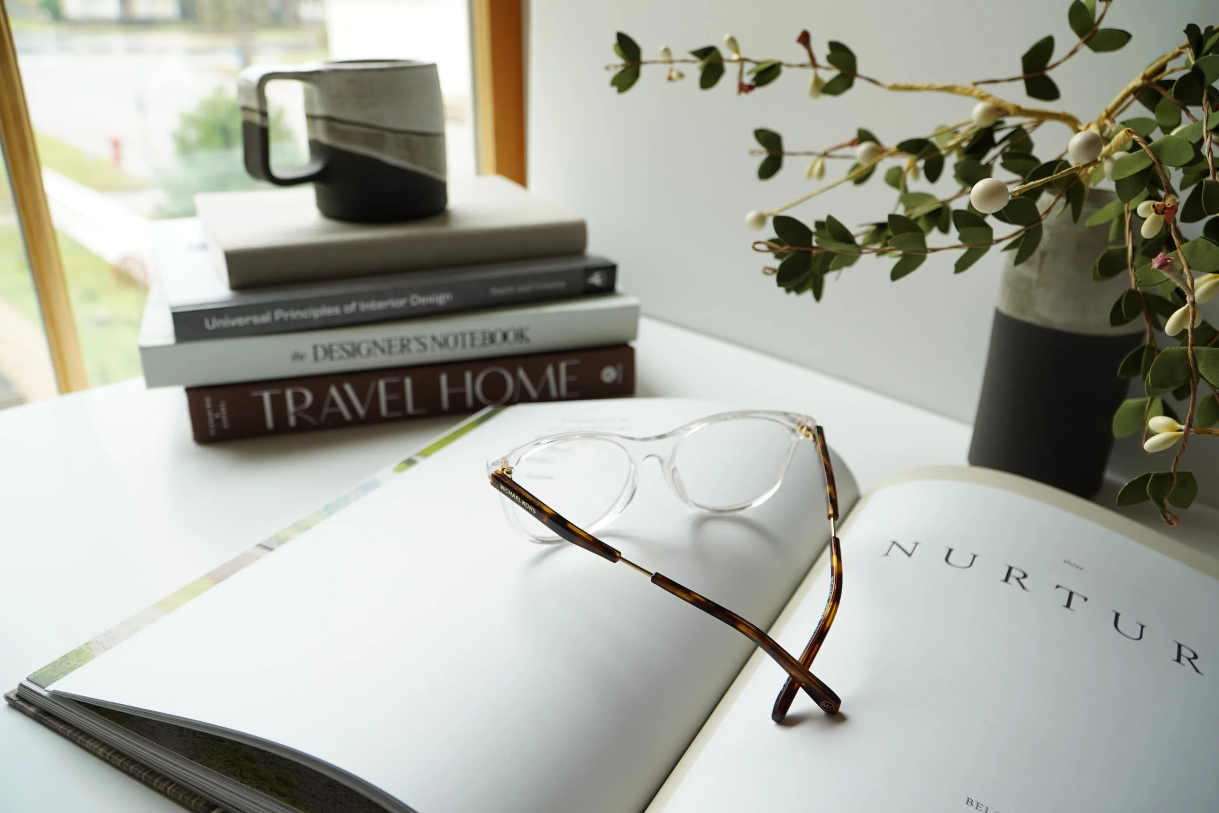 A white desk with an open book titled 'NURTURE', a pair of clear eyeglasses with tortoise shell temples resting on the book, a stack of books including 'Universal Principles of Interior Design', 'The Designer's Notebook', and 'Travel Home', a black and beige mug, and a potted plant with green leaves and white berries near a window.