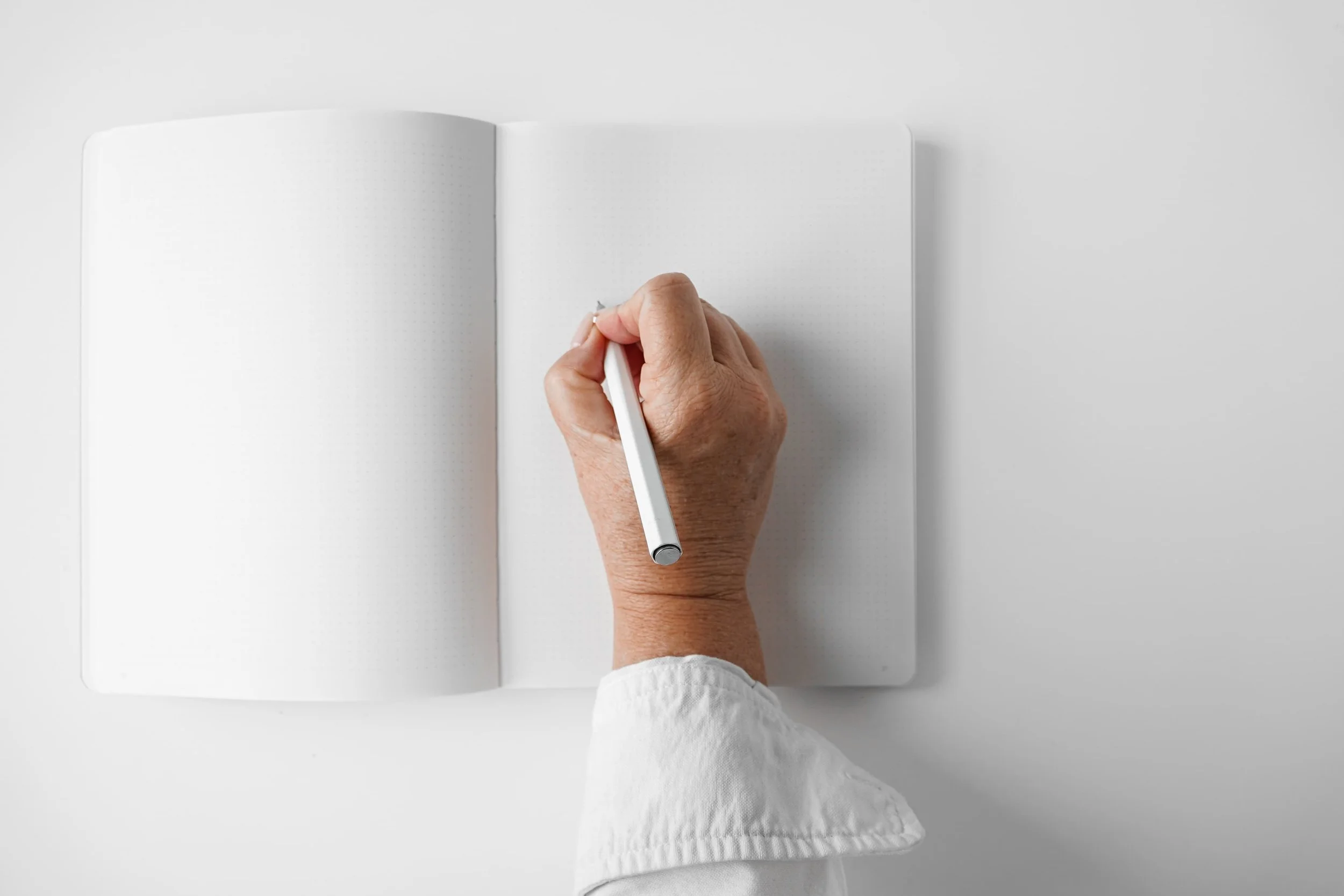 A person writing in a notebook with a white pen on a white desk.