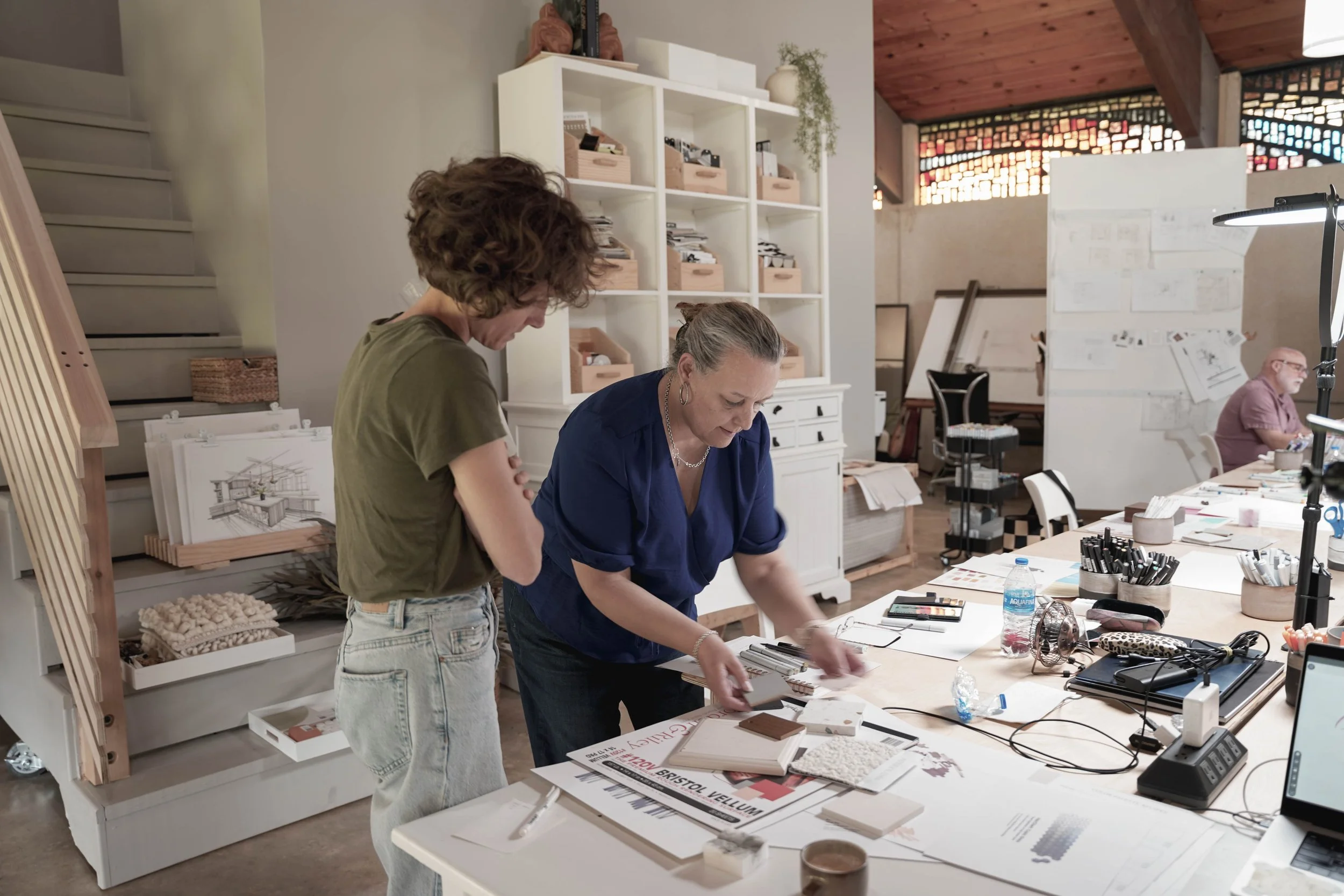 Two women are working together on a design project in a well-lit, modern workspace with shelves, drawings, and design materials on the table.
