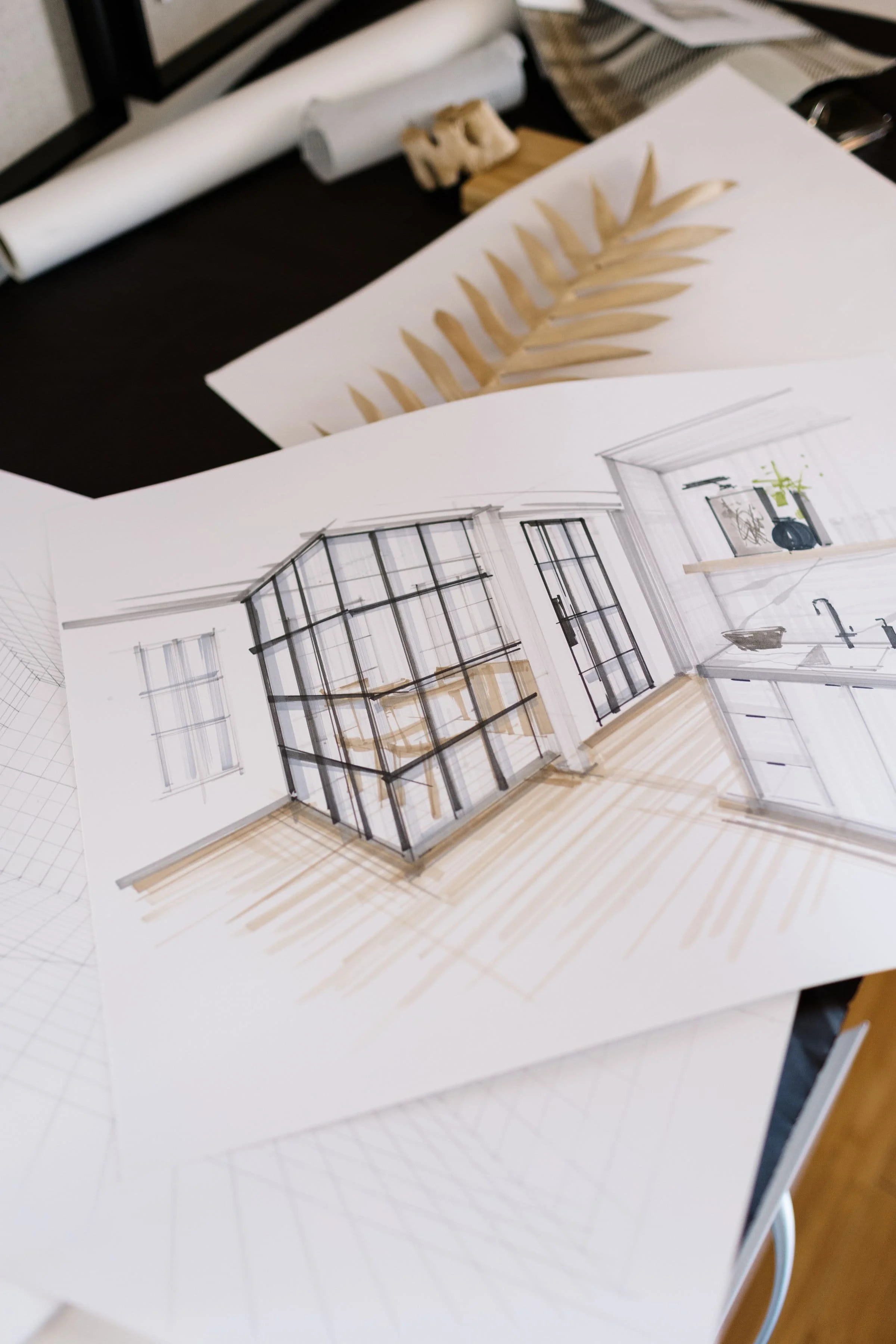 A detailed architectural sketch of a kitchen design with a glass cabinet, black framing, and wooden accents, placed on a workspace table with other sketches and design materials around.