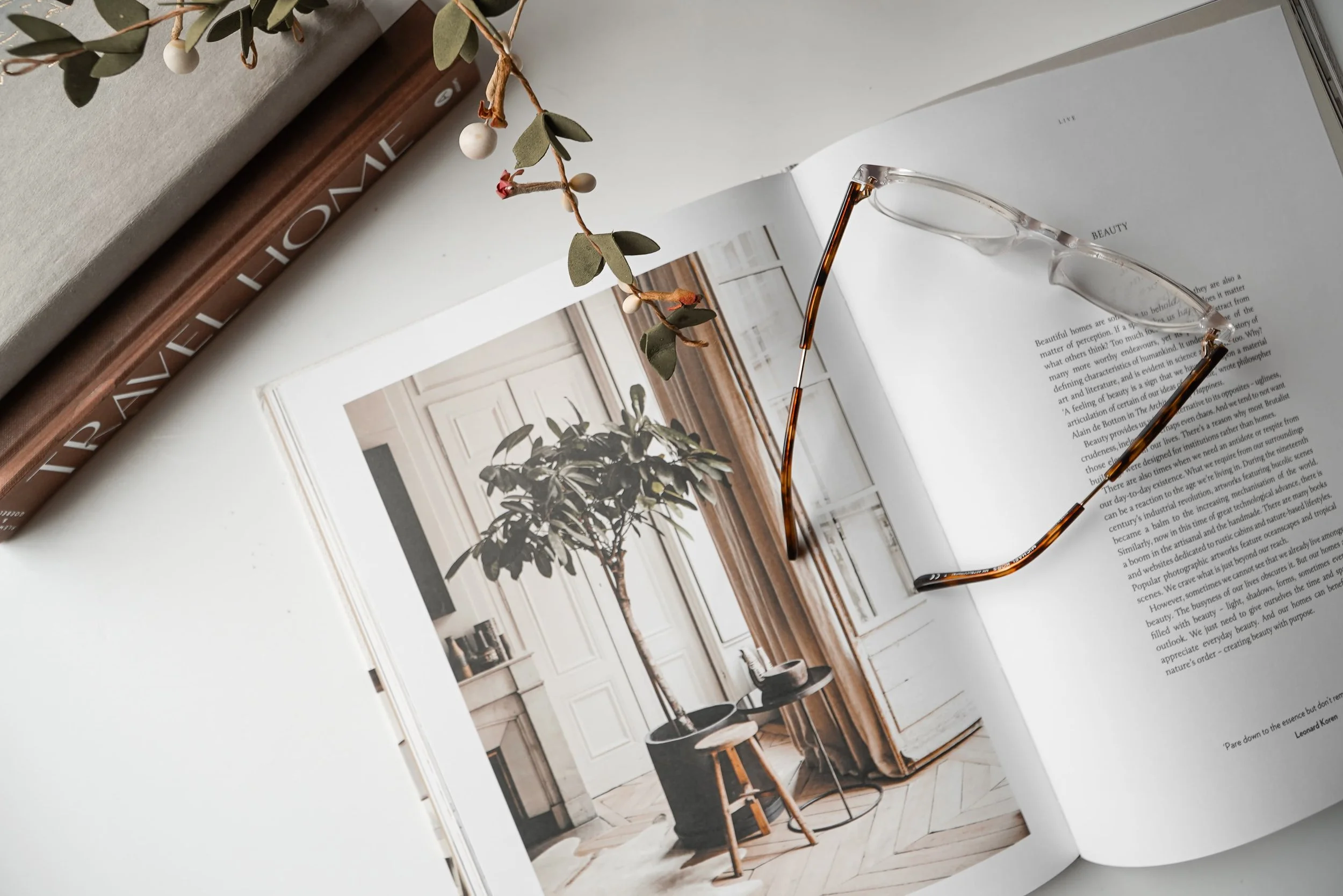 An open magazine displaying a photograph of an elegant interior with a tall potted plant, a chair, and panel doors. A pair of tortoise shell glasses rests on the page.