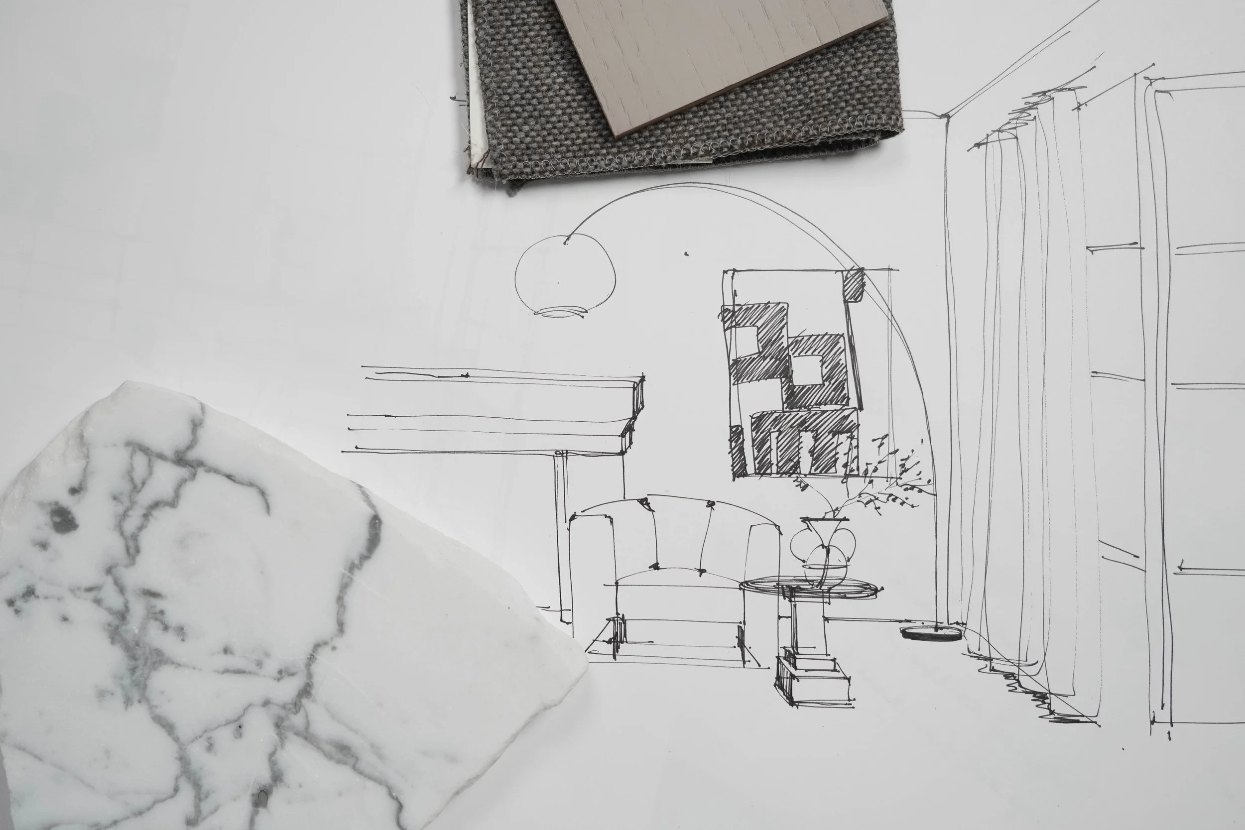 A design sketch of a living room layout with furniture, a hanging lamp, and floor-to-ceiling windows. A grained sample, a dark textile, and a wooden plank are placed on the sketch.