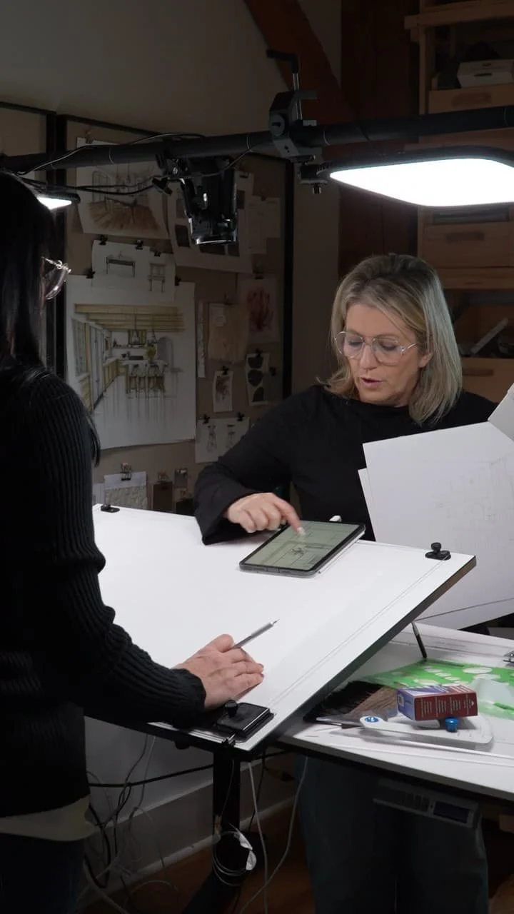 Two women working in a photography or art studio, with one using a tablet and the other holding a pencil, under studio lighting, surrounded by art supplies and equipment.