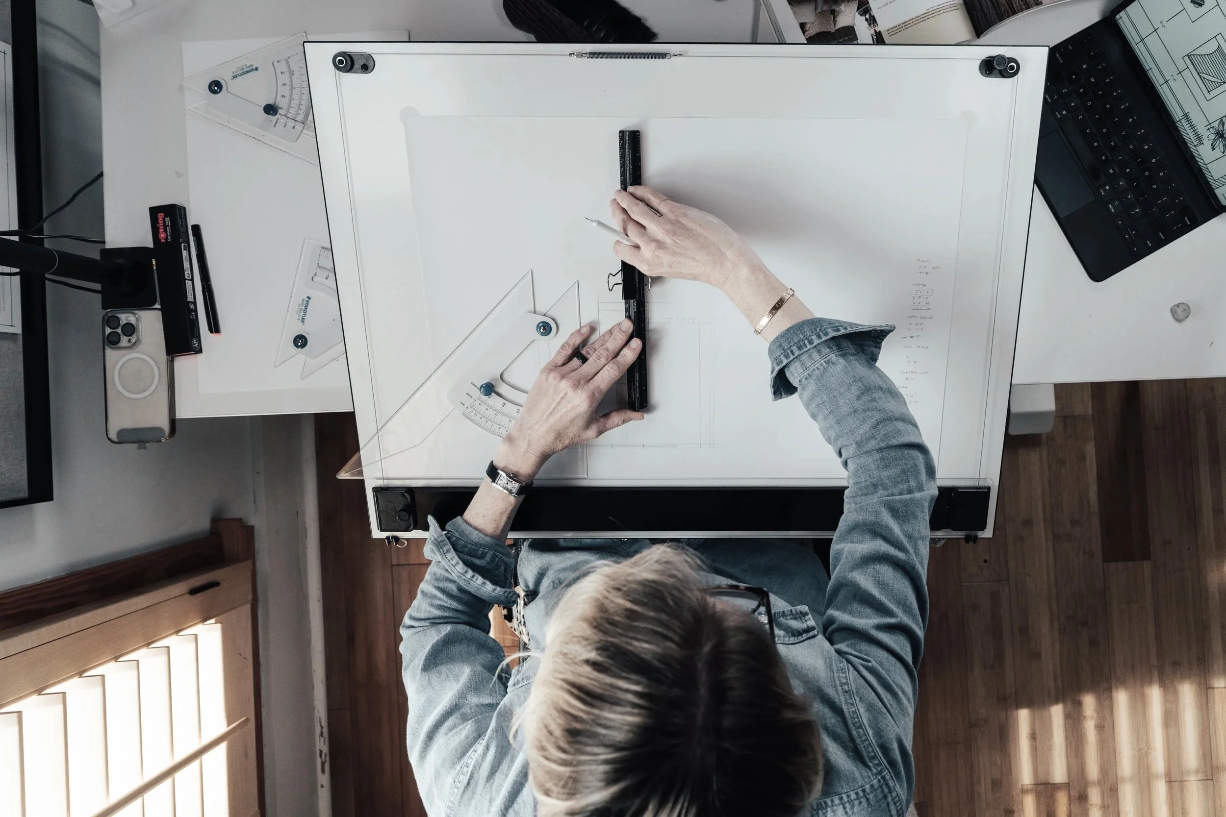 A person is working on a drafting table with tools like a ruler and triangle, surrounded by architectural drawings and a laptop, viewed from above.