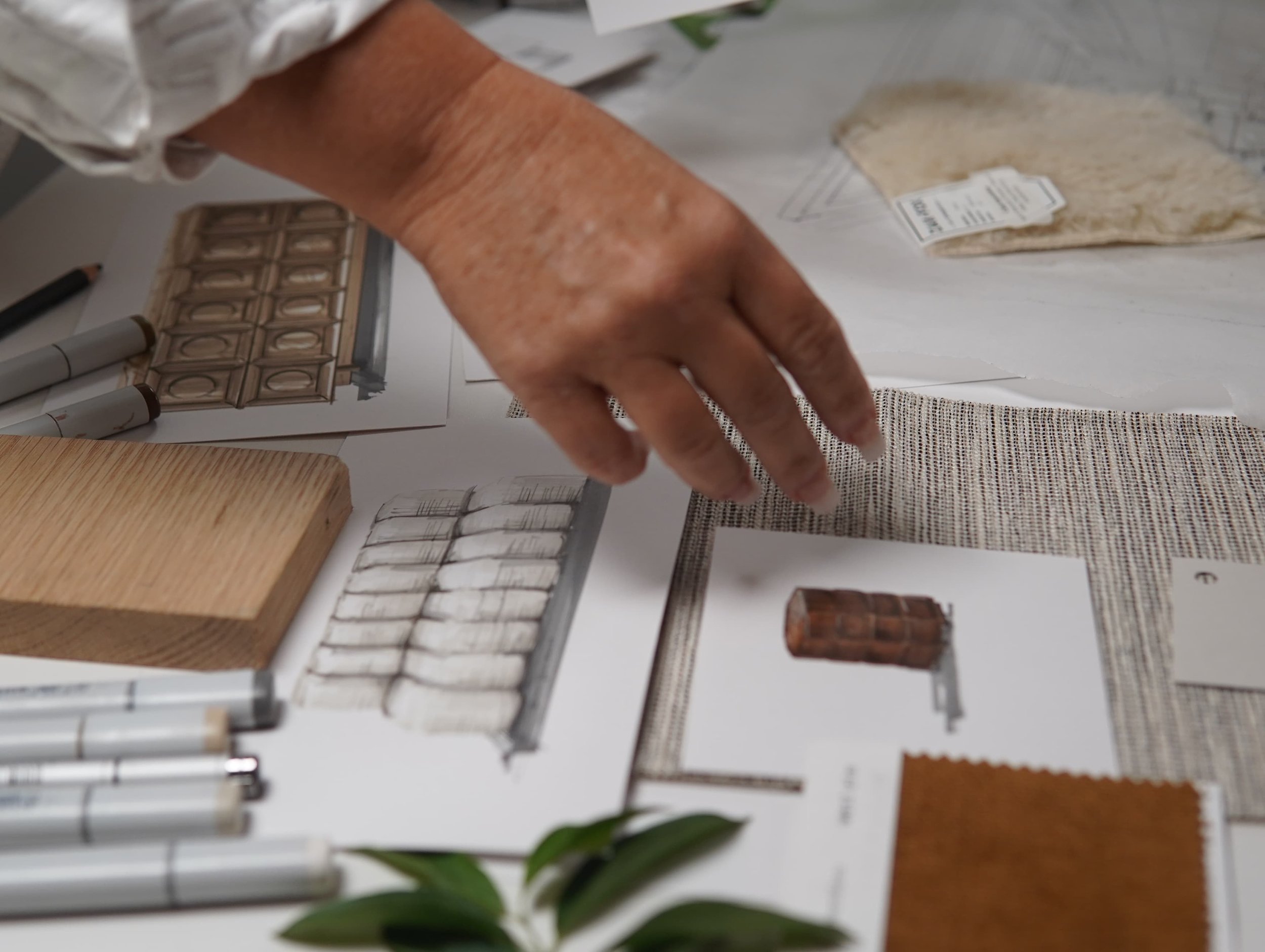 Hand of a person with aged skin pointing at fabric samples on a table, surrounded by design documents, fabric swatches, and drawing tools.