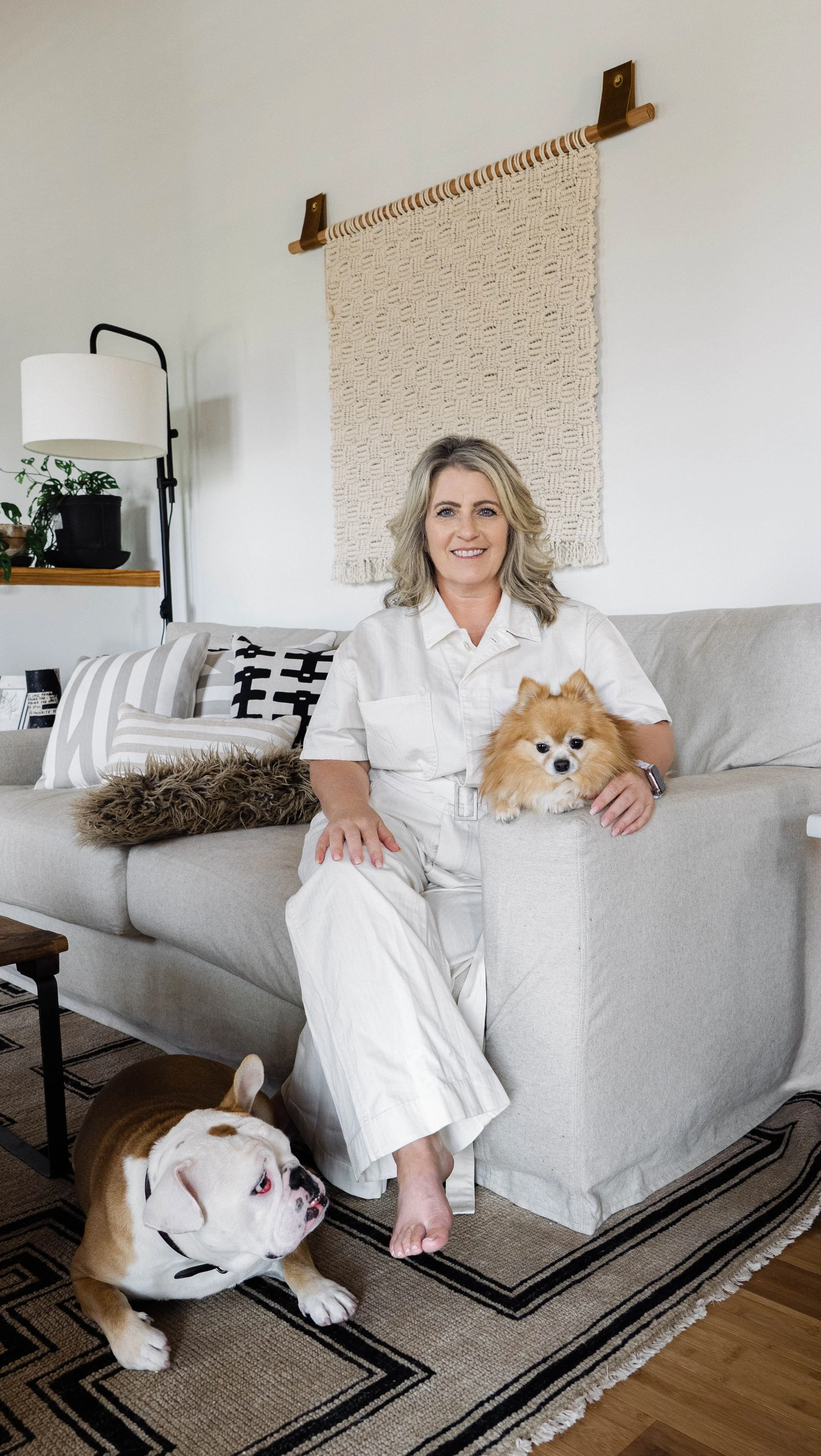 A woman sitting on a beige sofa with two dogs, a Pomeranian on the armrest and a Bulldog on the floor, in a cozy living room with a woven wall hanging behind her.
