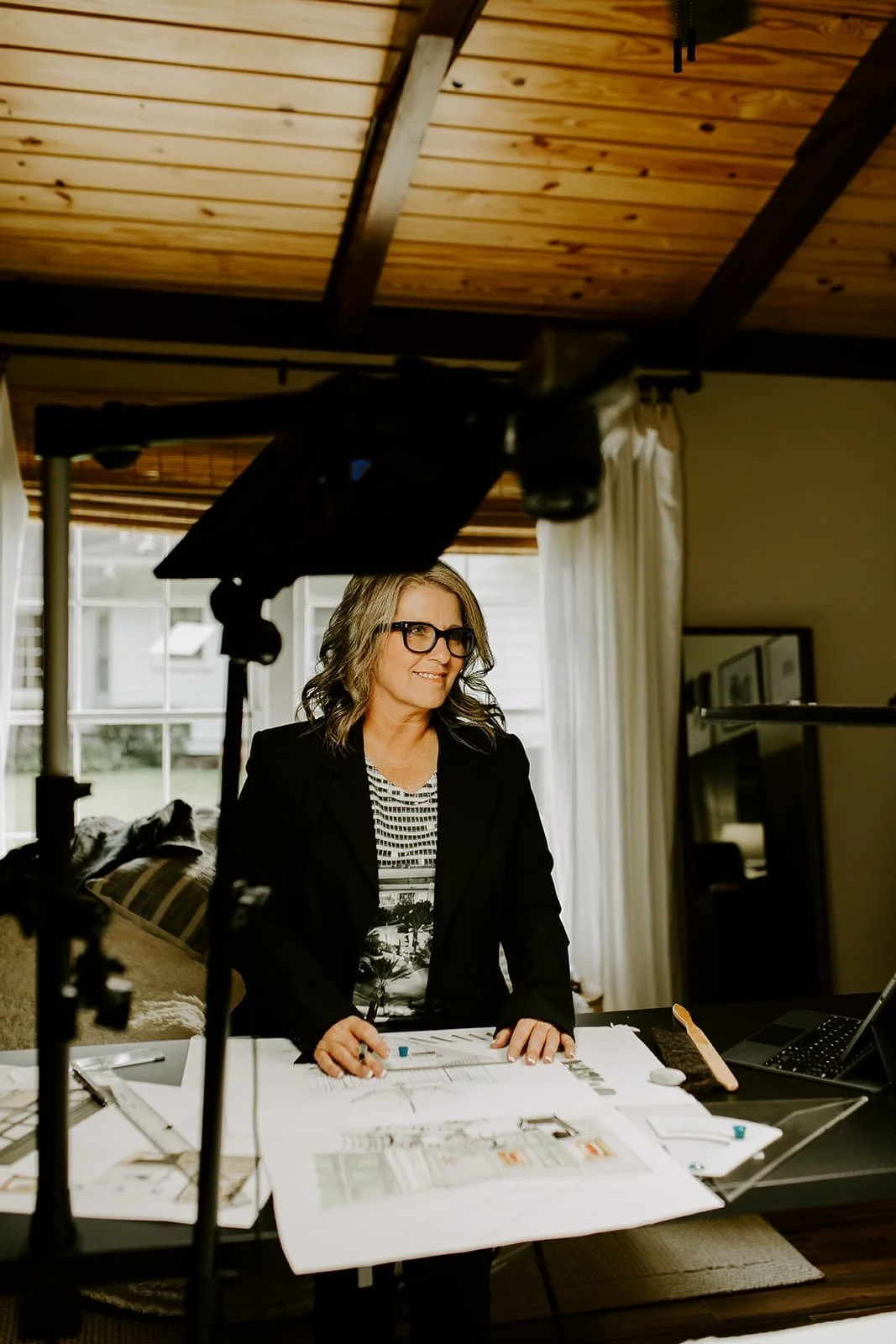 A woman with glasses and wavy gray hair standing at a table with architectural plans, smiling and looking to her right, in a room with a wooden ceiling and a large window in the background.