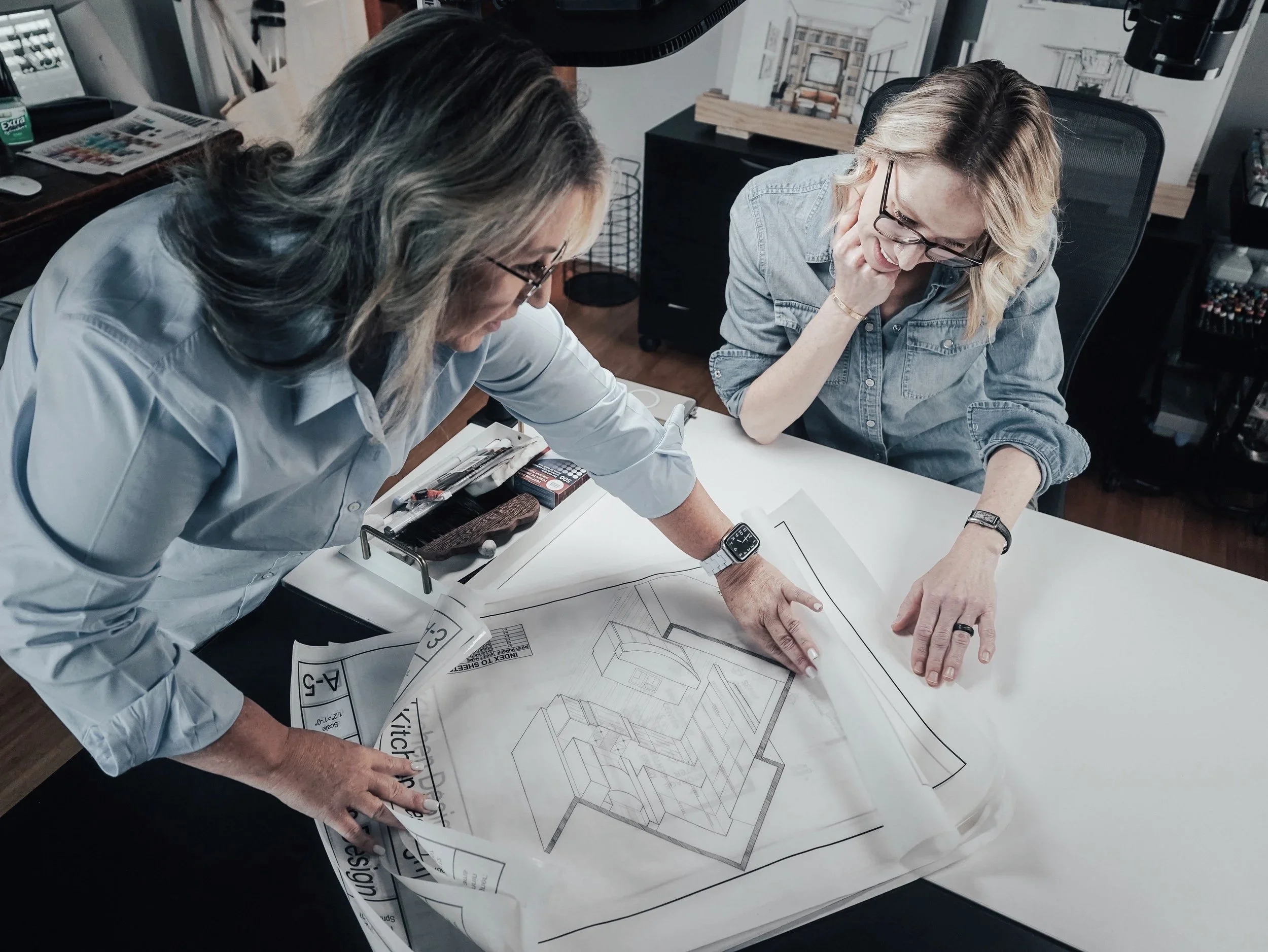 Two women, both wearing glasses, review an architectural blueprint on a white table in an office. One woman is pointing at the blueprint while the other looks on, with various office items and artwork in the background.