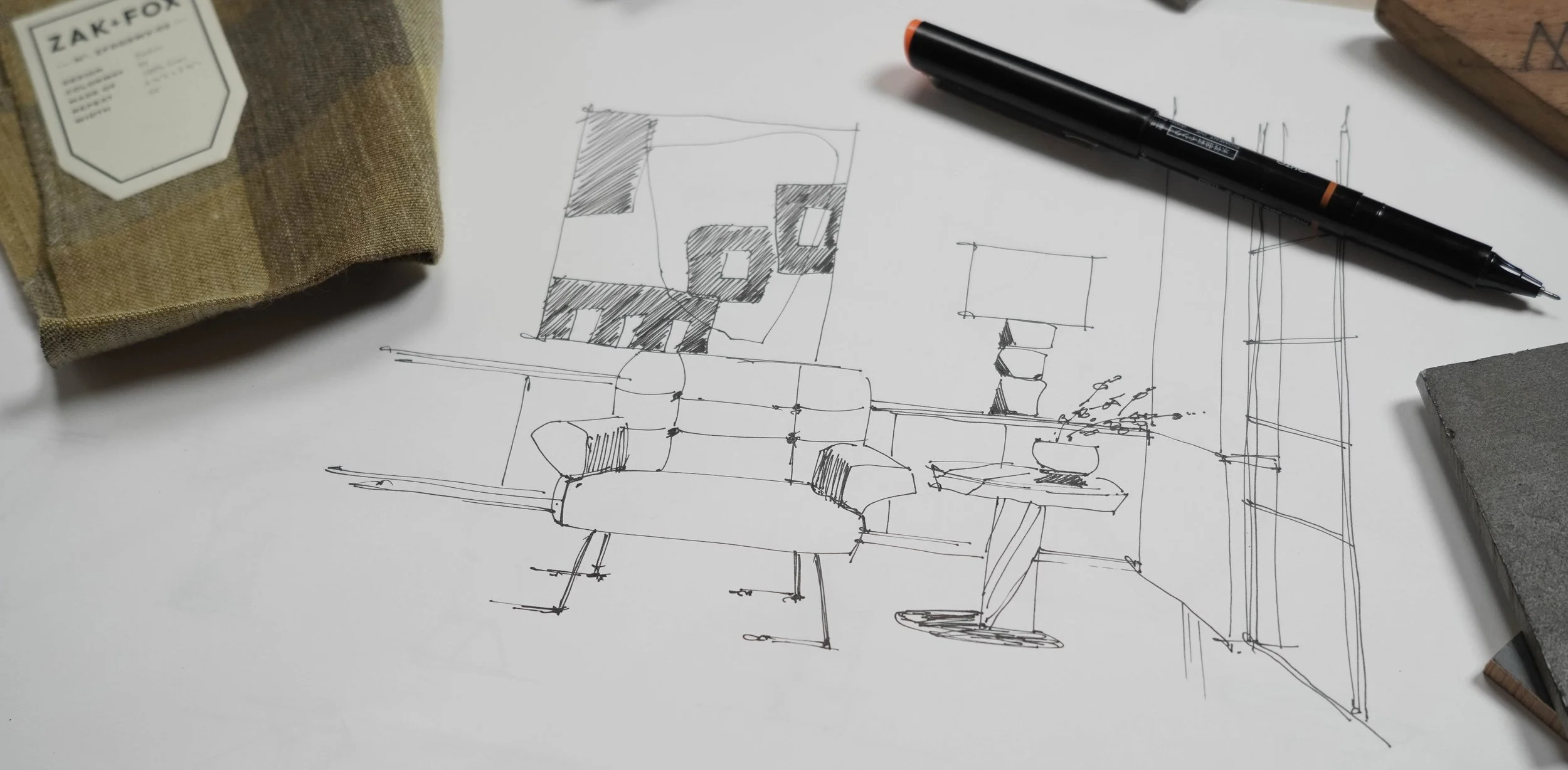 Sketch of a living room interior with furniture, behind which are a potted plant, coffee table, and shelves, drawn on a piece of paper on a white desk. Items on the desk include a black marker, a fabric-covered object on the left, and a gray rectangular object on the right.