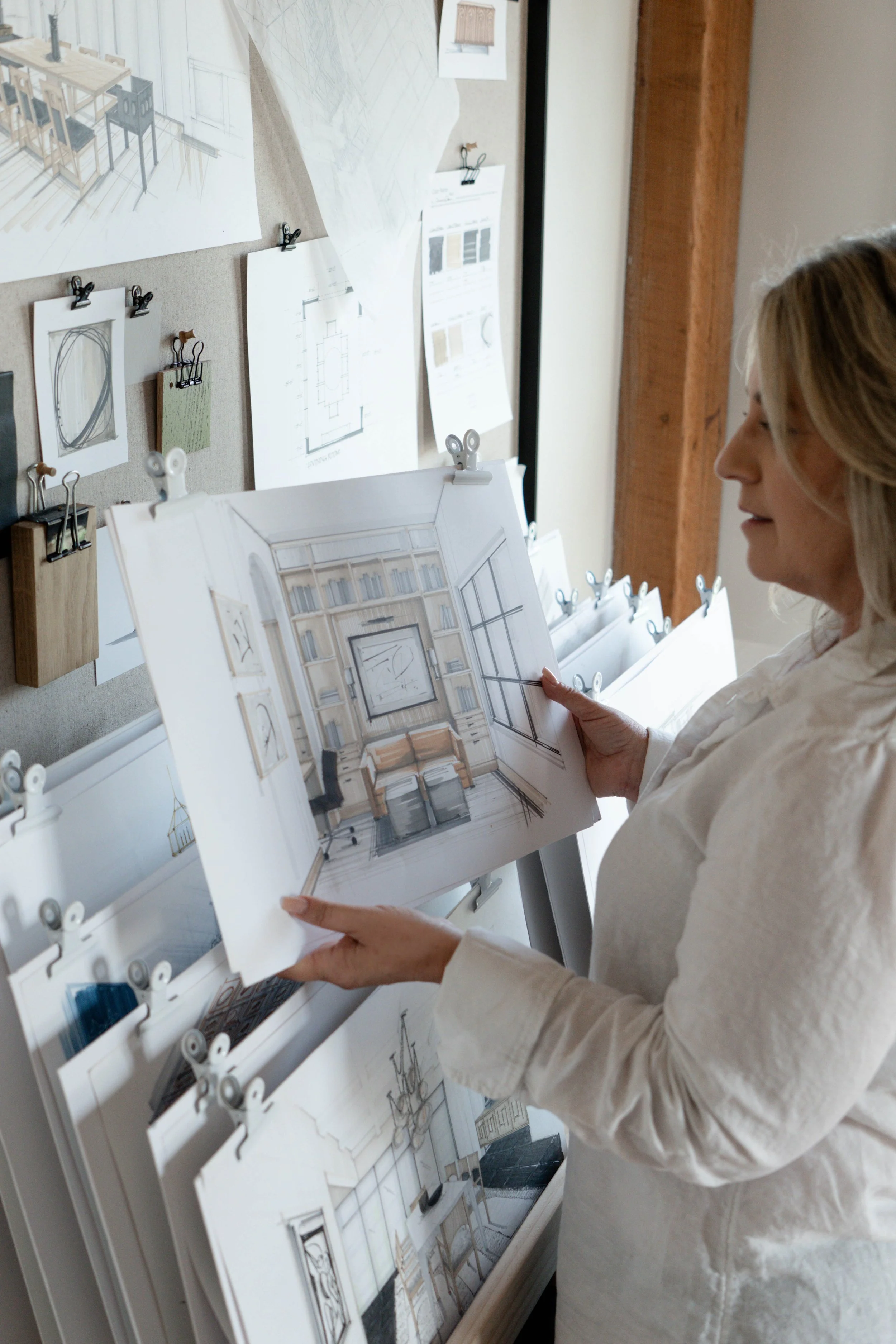 Woman examining interior design sketches and mood boards pinned to a conference room wall.