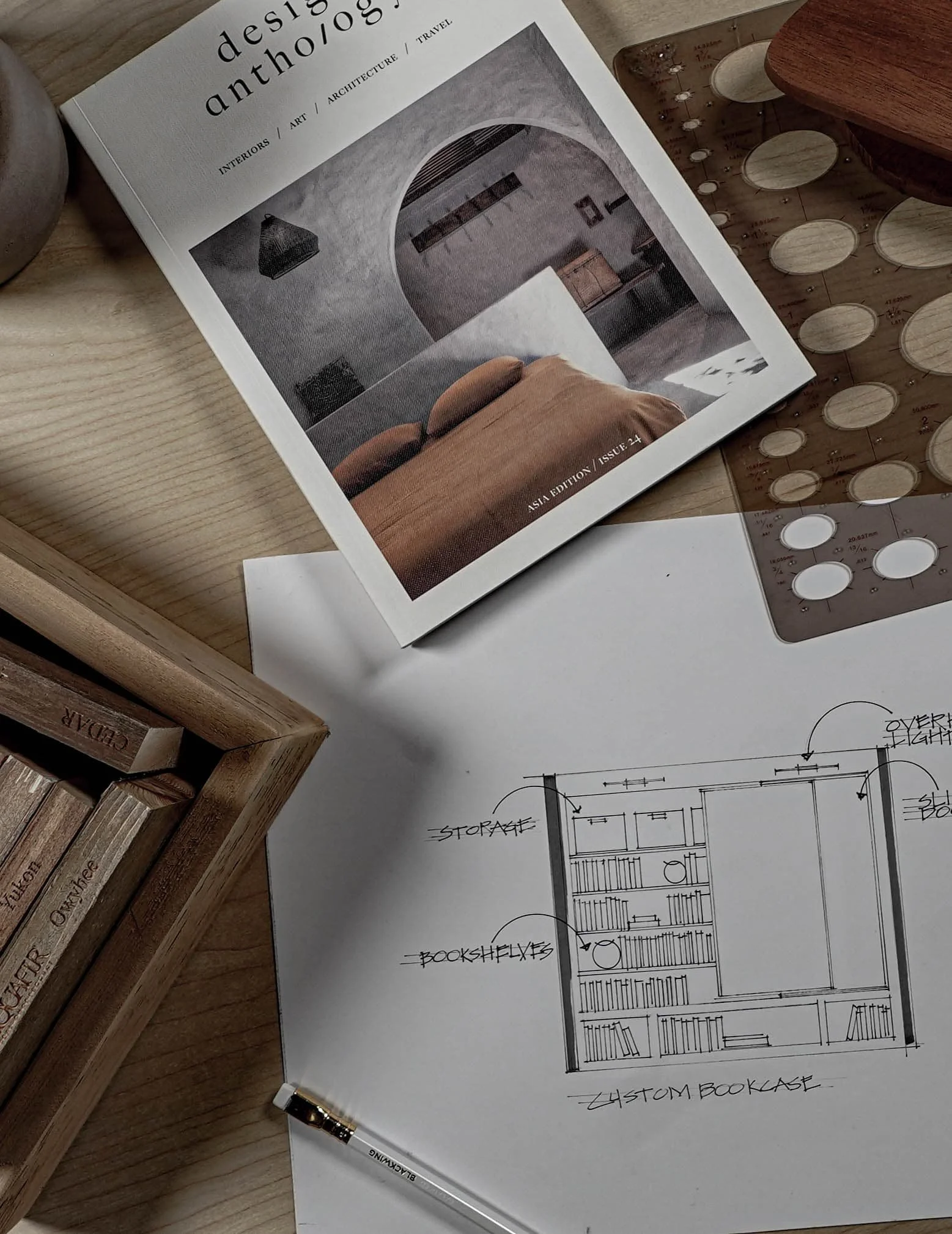 A black and white architectural drawing of a custom bookcase layout on a piece of paper, surrounded by a white pen, a wooden bowl, a magazine about design, and a wooden box on a wooden surface.