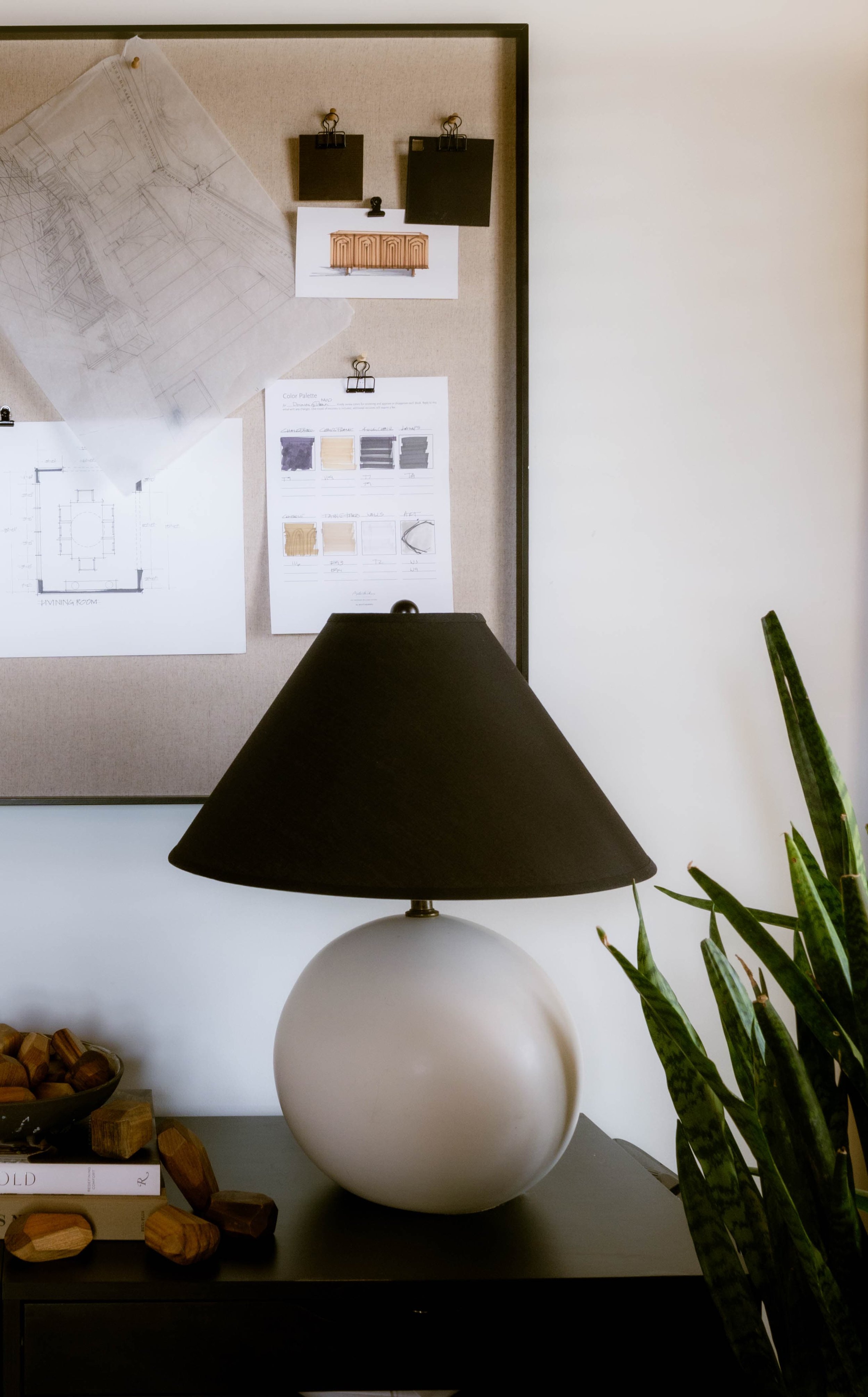 A black and white table lamp with a spherical base and black shade on a black surface. To the right, there is a tall green plant with long leaves. Behind the lamp, a cork bulletin board displays architectural plans, color palettes, and swatches, with some papers and images clipped to it.