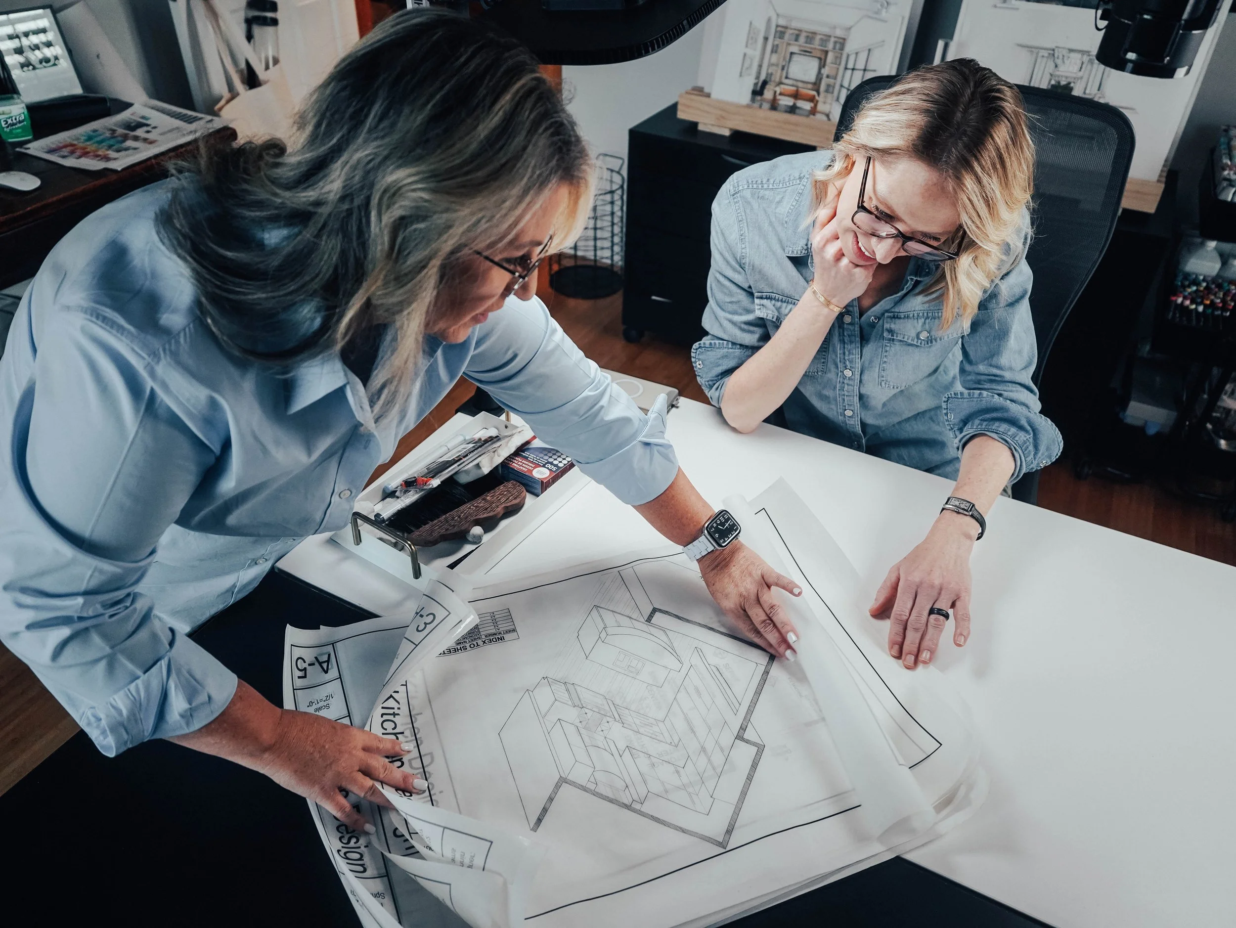 Two women examining architectural blueprints on a table in an office setting.