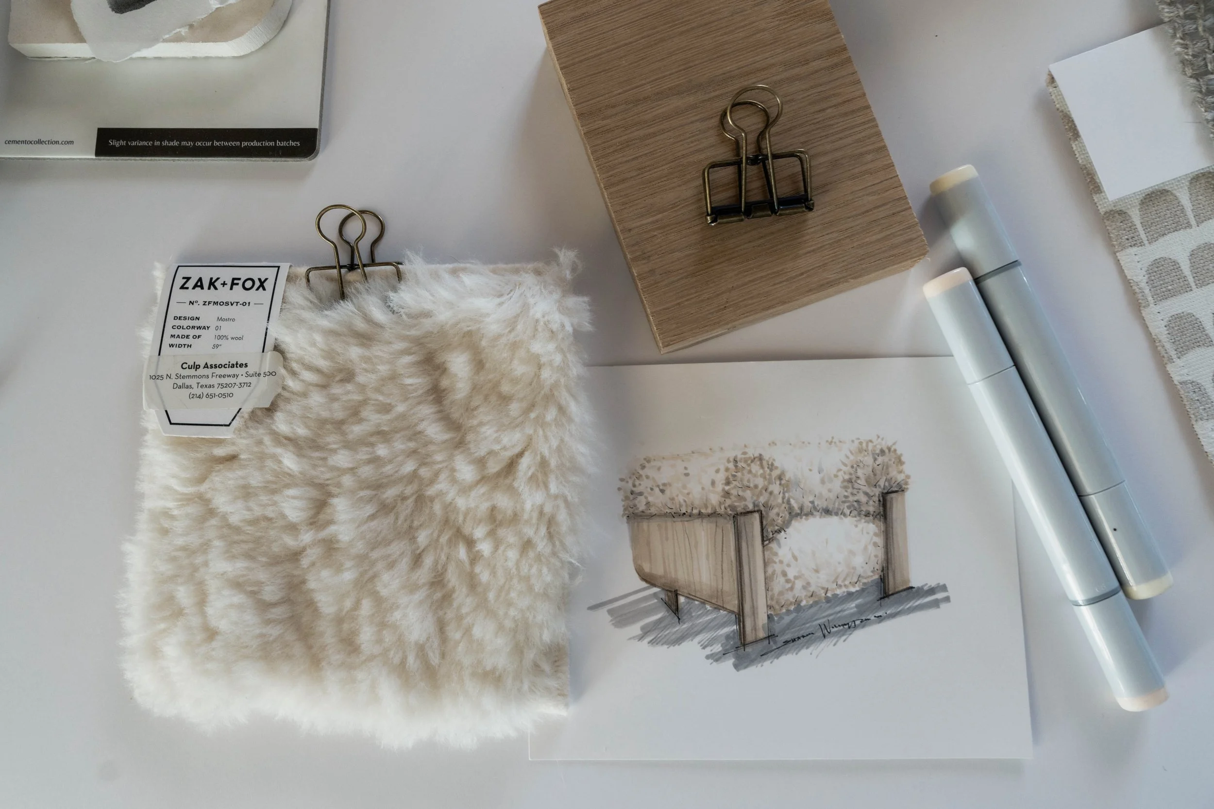 Sample swatch of cream-colored faux fur fabric with a hanging label, a small wooden block with metal clips, a color sketch of a cream textured bathtub, two white markers, a piece of fabric with semi-circular patterned cutouts, and a paint swatch card.