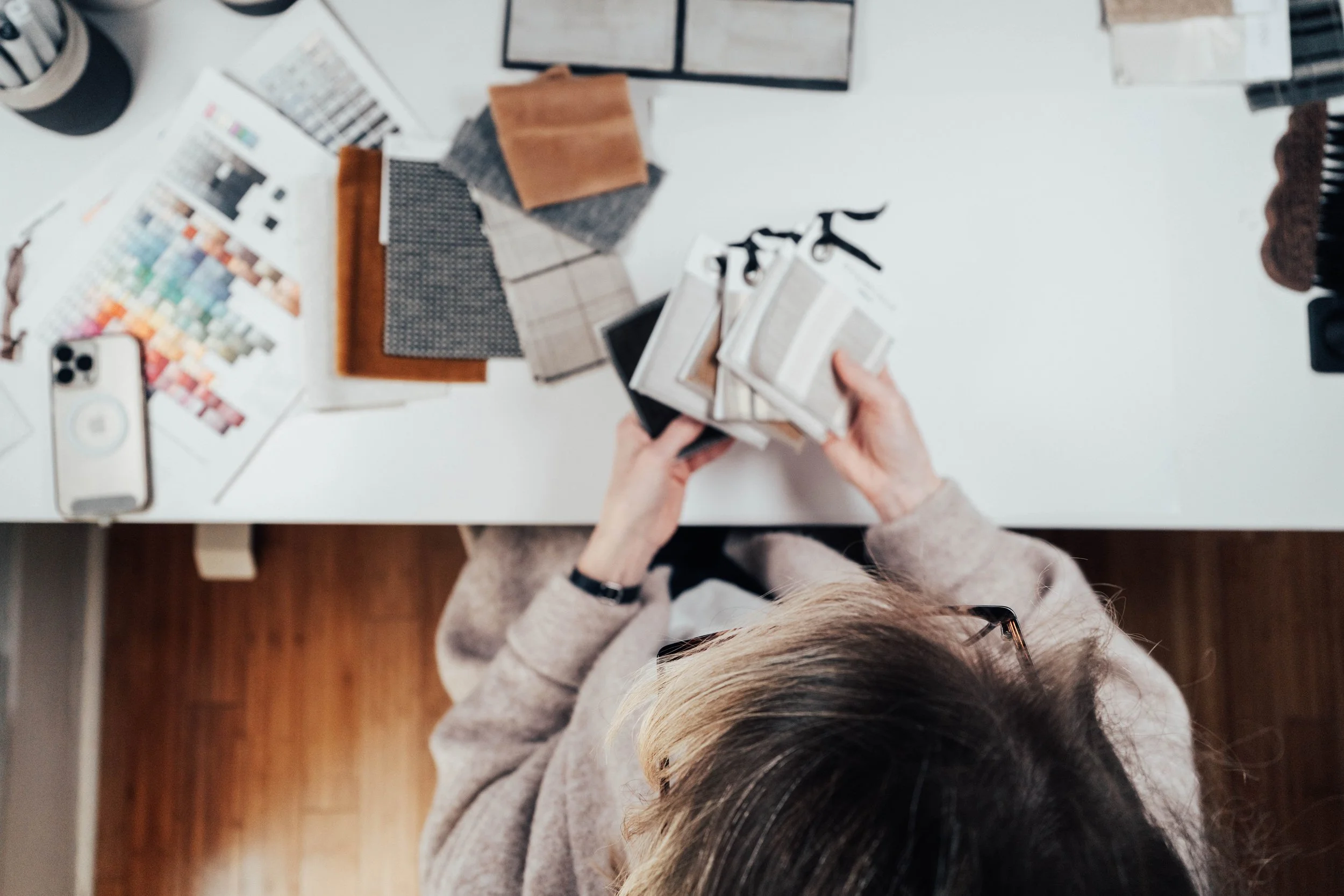 Top-down view of a woman organizing fabric swatches on a white table. The table has various color swatches, fabric samples, a smartphone, color palette, and measurement tools.