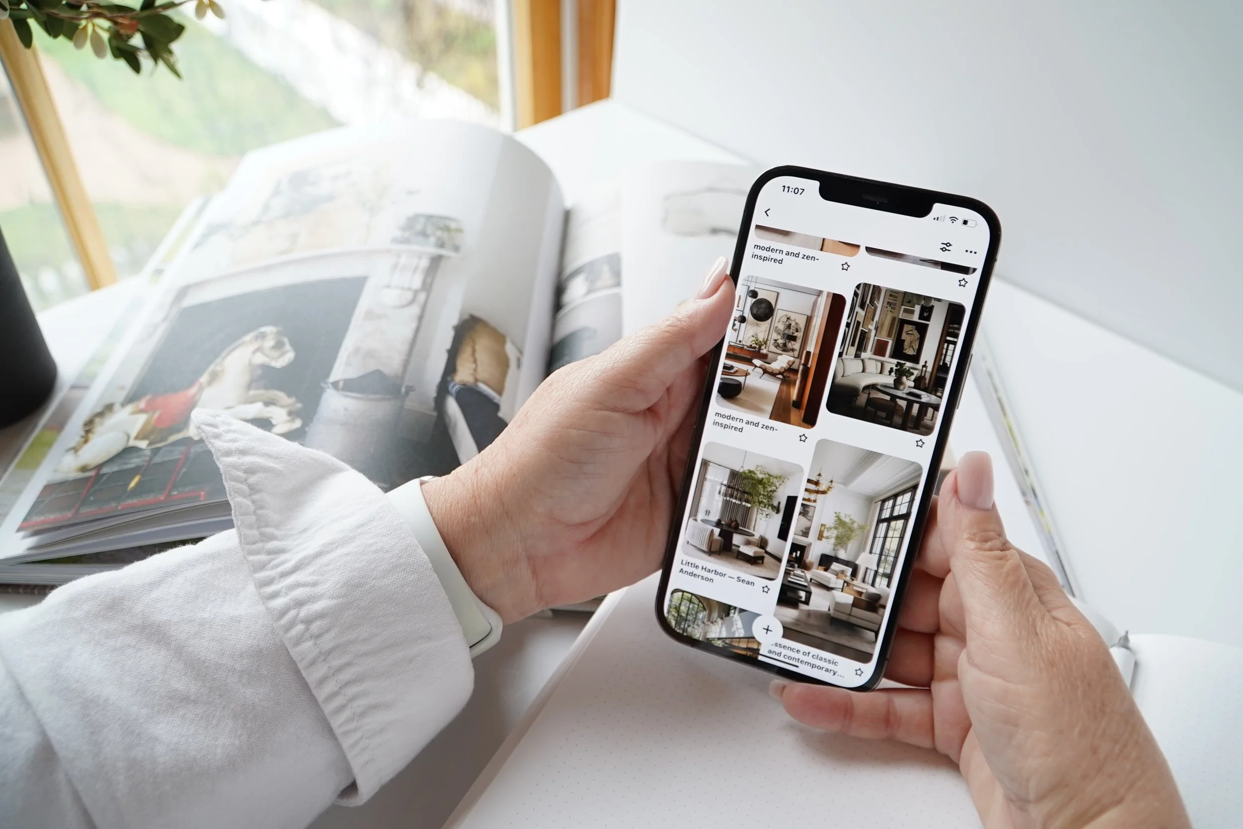 Person holding a smartphone displaying interior design images, with an open magazine on a white desk and a window in the background.