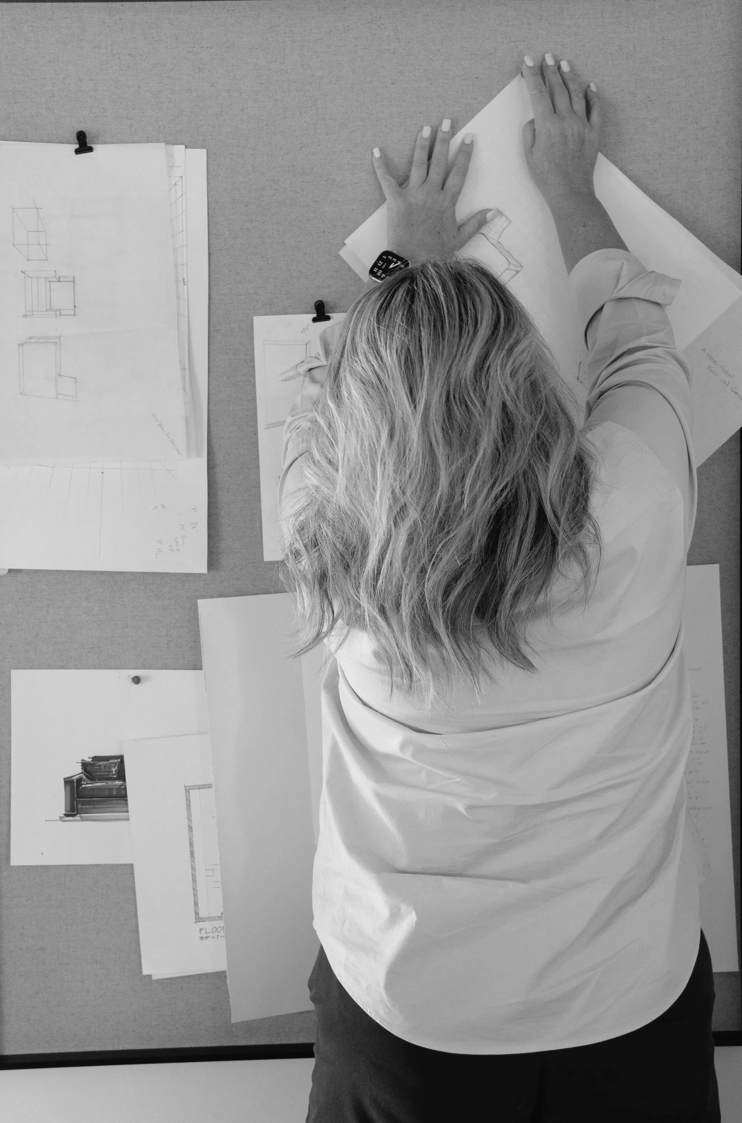 A woman with wavy hair is working on architectural drawings at a desk, with sketches and plans laid out around her.