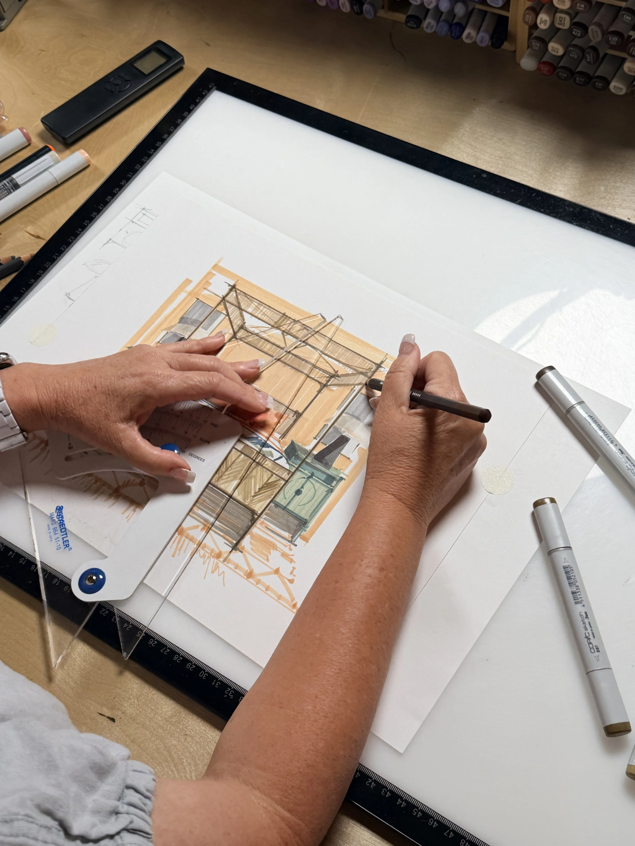 Person working on architectural or interior design sketches, using markers and pencils, with a printed design and technical drawing on a drafting board.