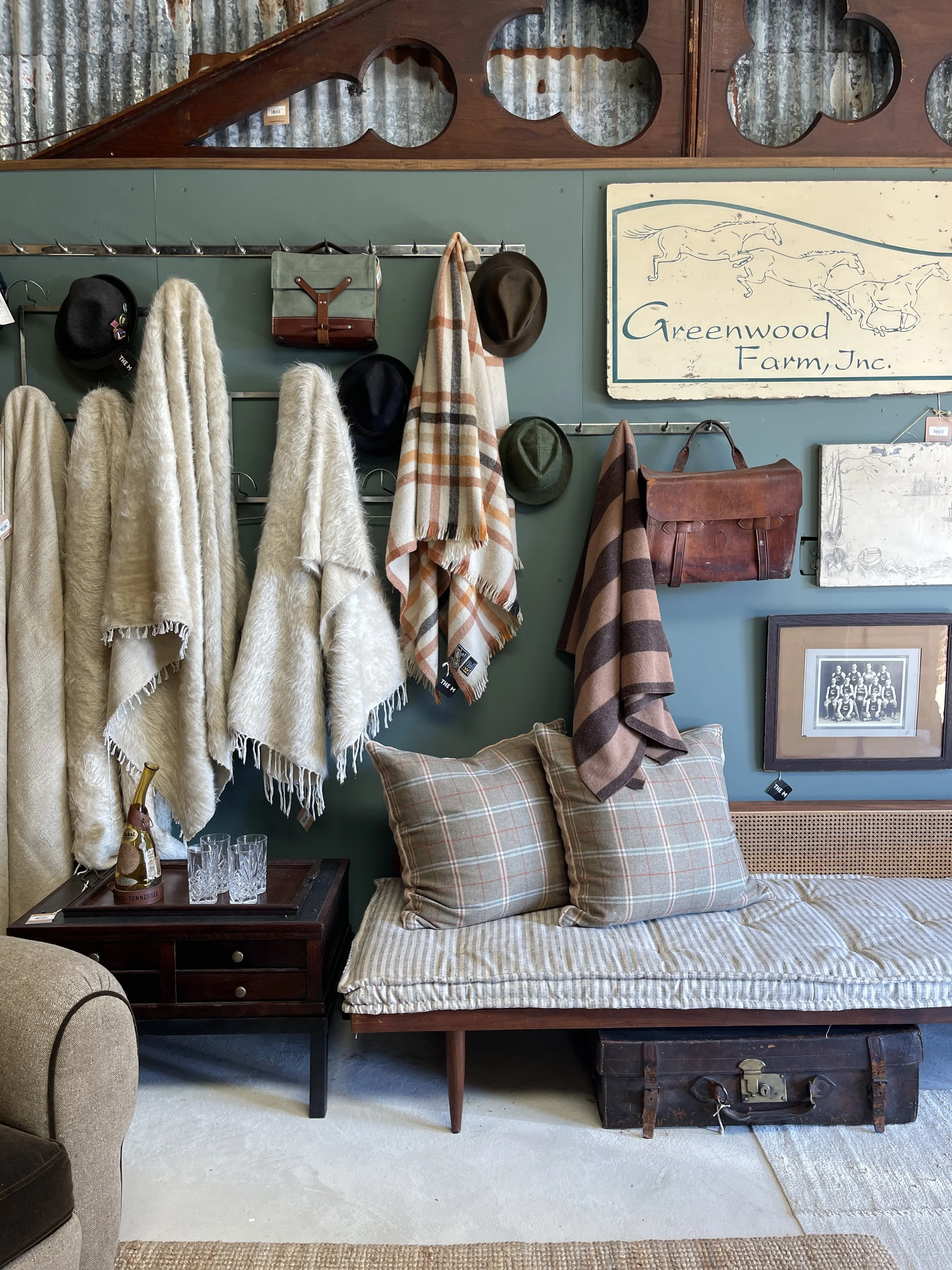 Display of vintage decor items such as hats, scarves, and a leather bag on a green wall. A sign reading 'Greenwood Farm Inc.' with illustrations of running horses. A bed with plaid pillows and blanket, a wooden bedside table with glasses and a bottle, and an old chest at the foot of the bed.