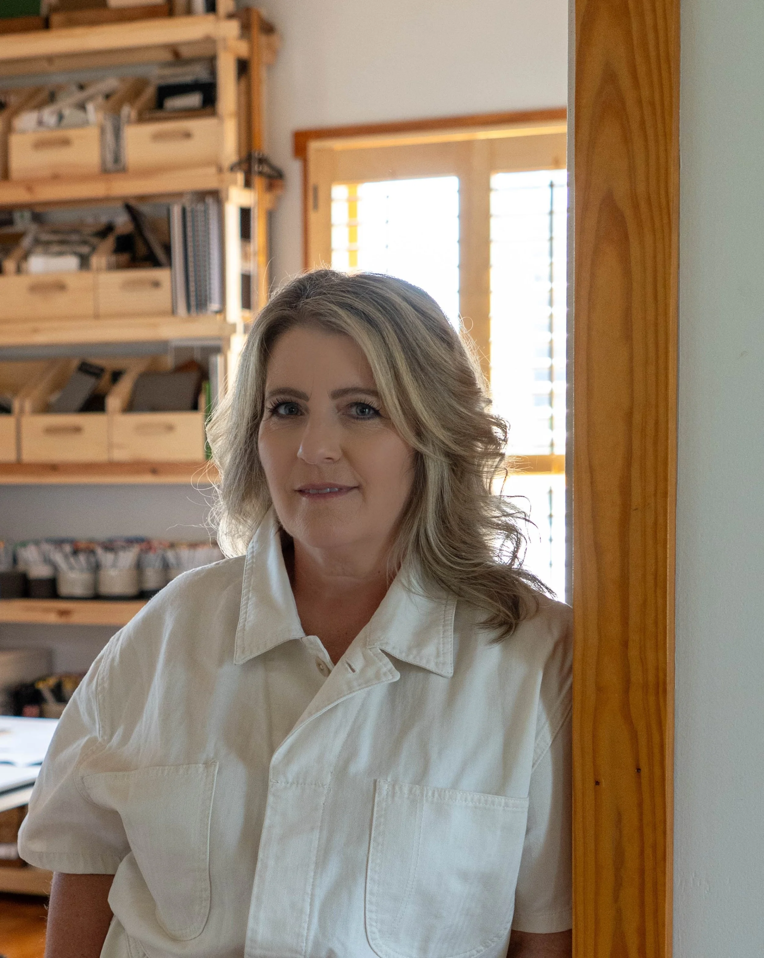 A woman with blonde hair wearing a light-colored button-up shirt, standing indoors near a wooden doorframe, with shelves of organized items and a window with blinds in the background.