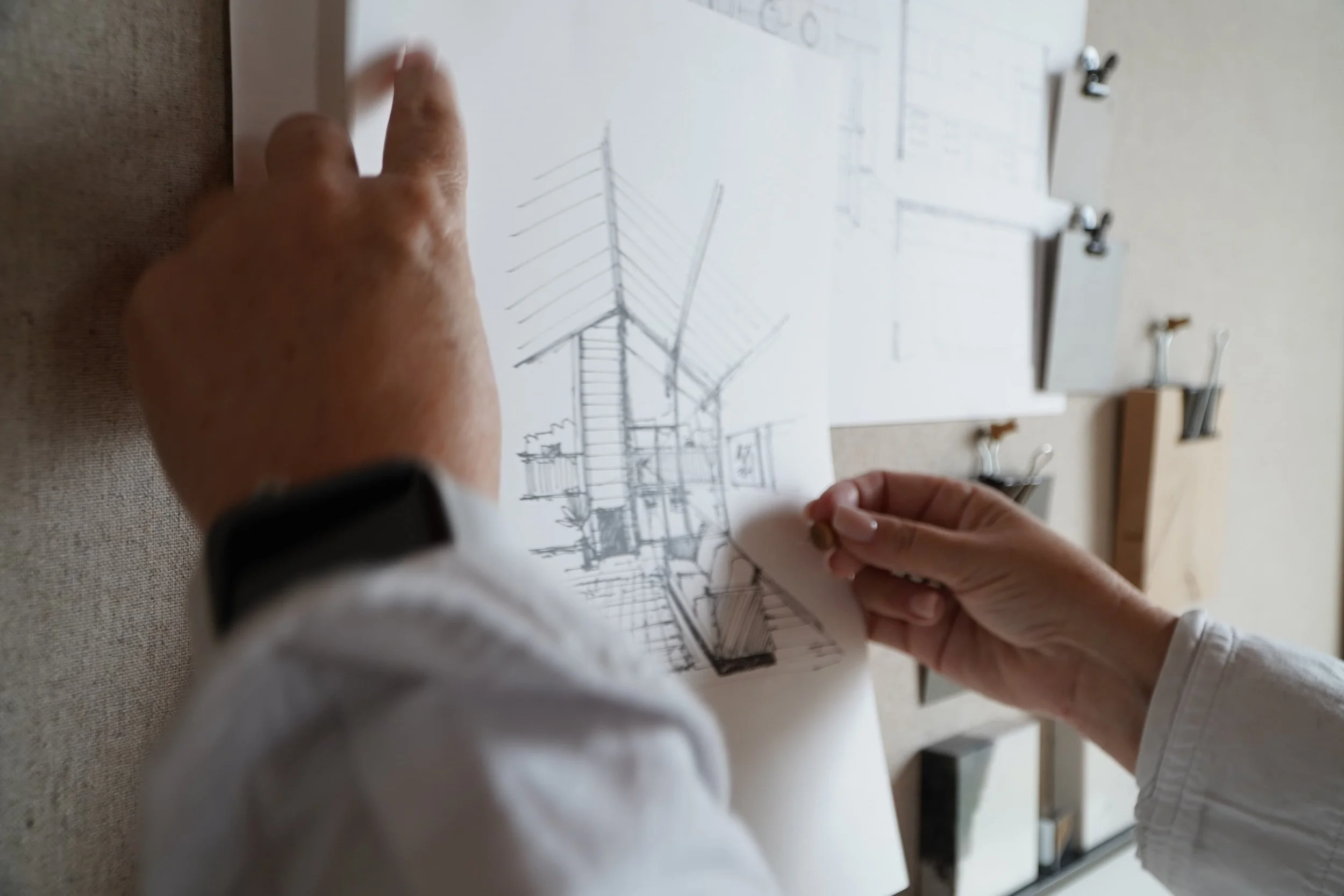 Person holding architectural sketch of a building, with drafting tools and design papers on a table.