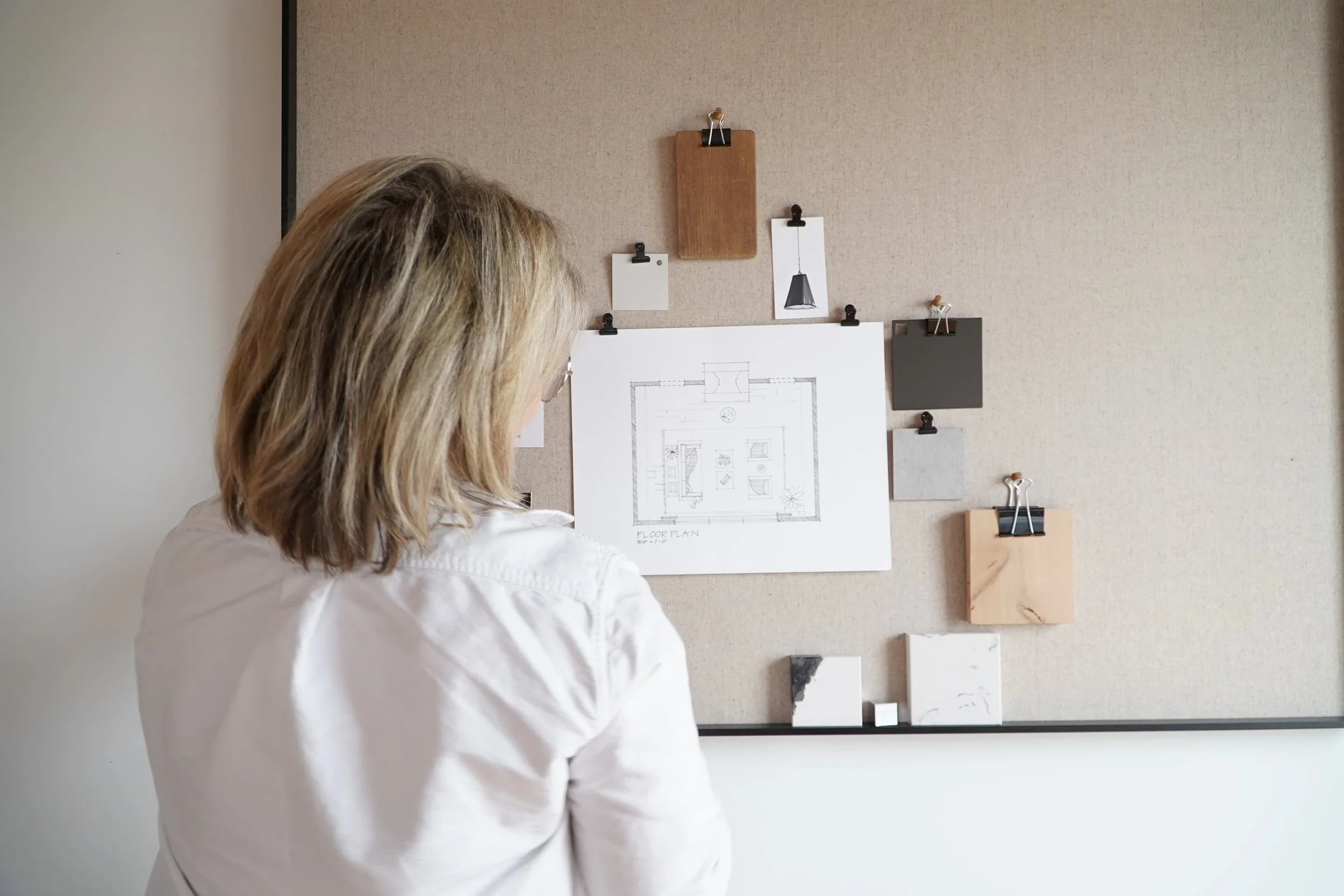 Woman with shoulder-length blonde hair in a white shirt looking at a corkboard wall with various pinned items, including a floor plan drawing, small pictures, and sample materials.