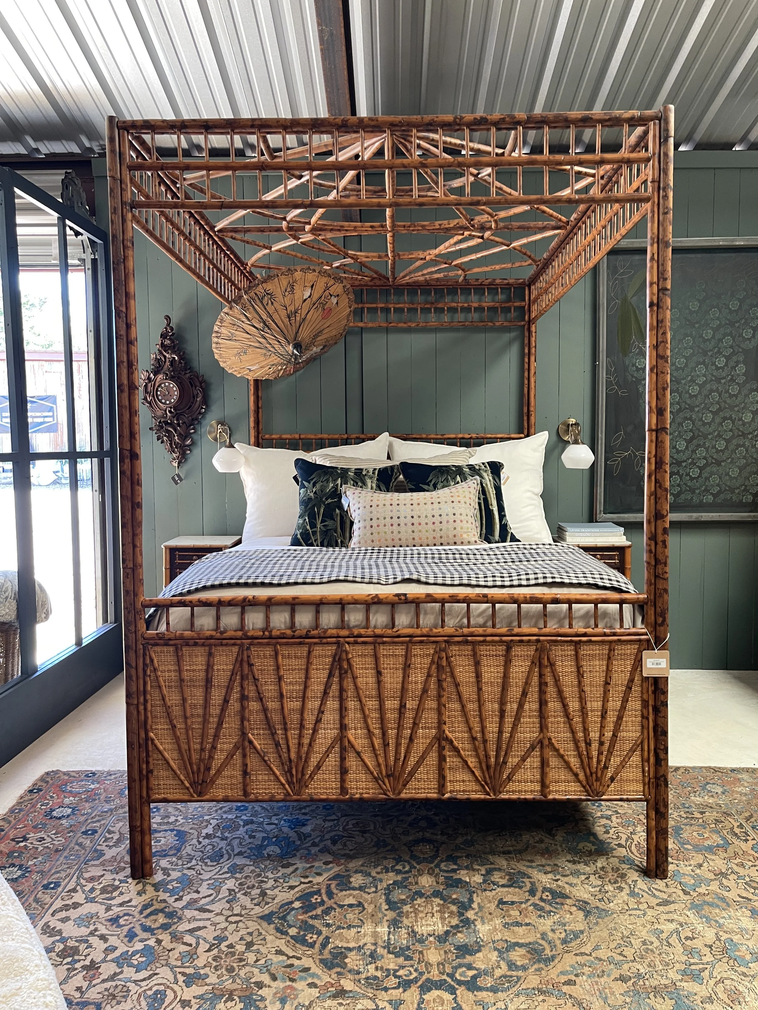 A bedroom with a large wooden canopy bed, featuring bamboo-style posts and a woven headboard, topped with decorative pillows and a gingham blanket, with a patterned area rug on the floor and a green paneled wall in the background.