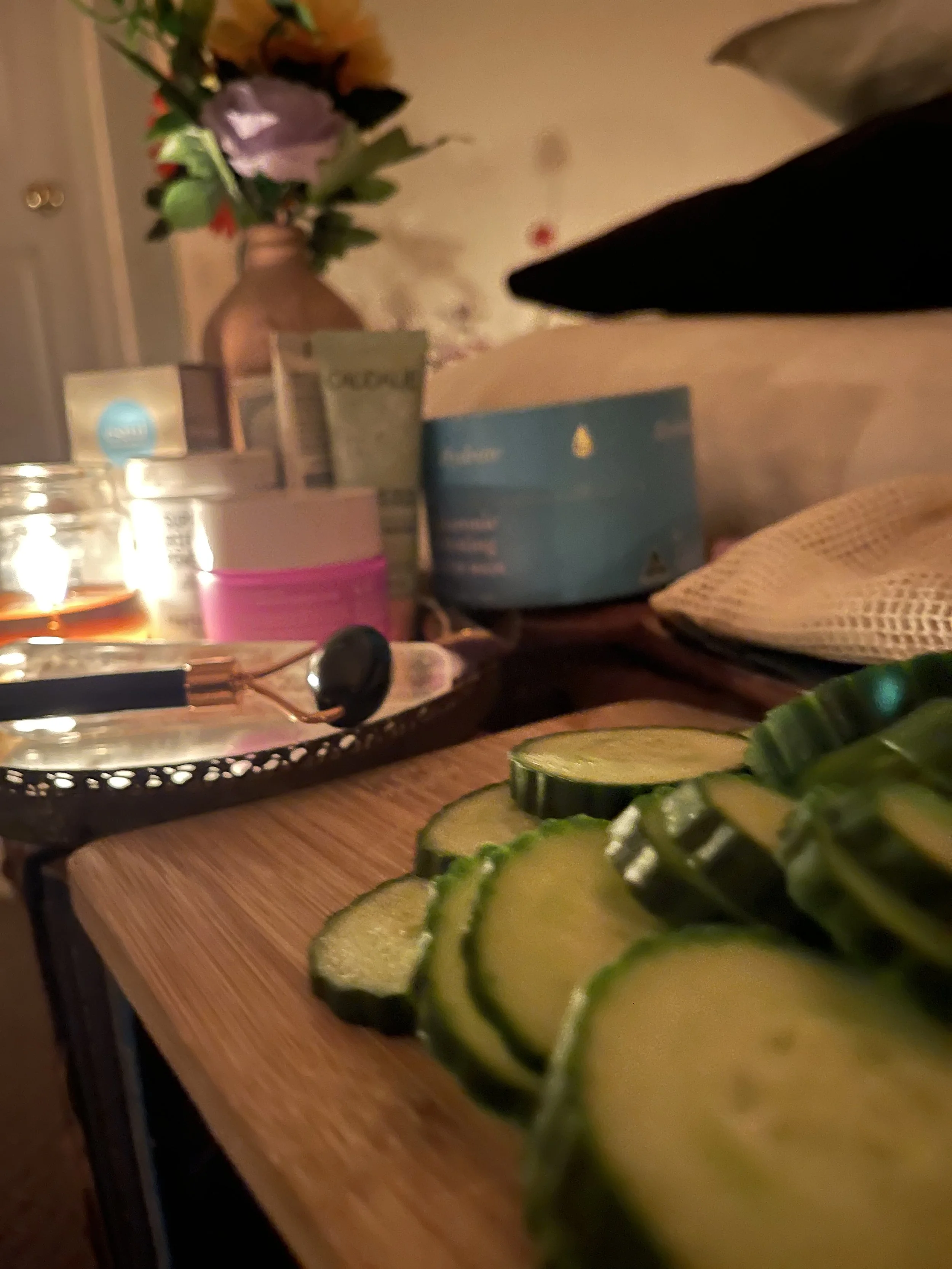 Sliced cucumbers on a wooden cutting board with skincare products, candles, and a flower arrangement in the background.
