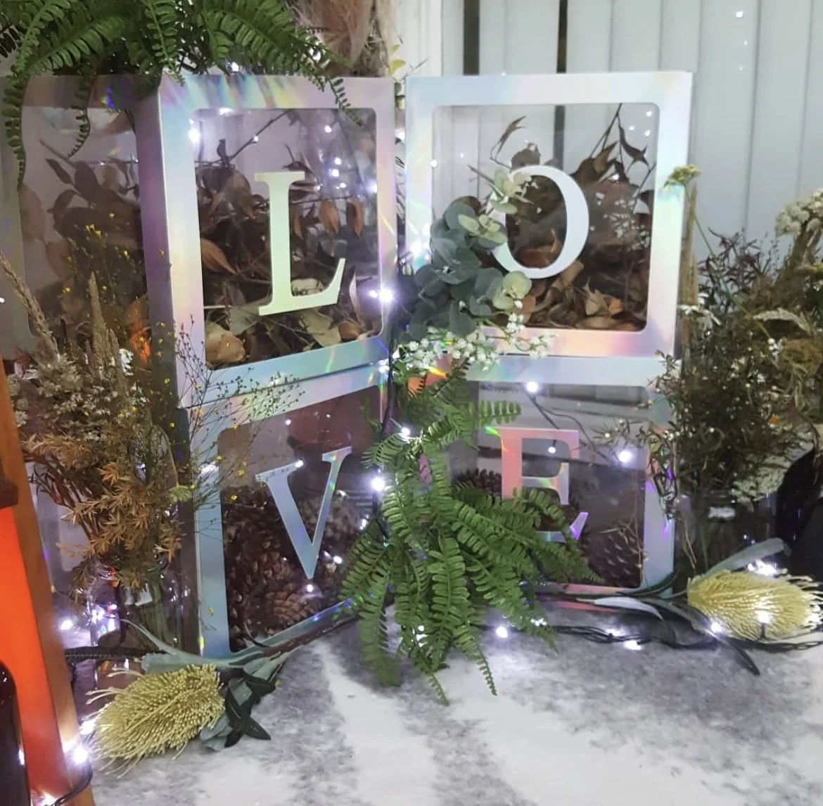 Decorative display of three glass boxes with the letters L, O, V, and E inside, surrounded by flowers, greenery, and string lights.