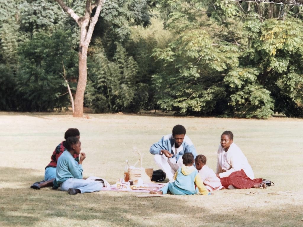 A group of six people, including children and adults, enjoying a picnic on a grassy field with trees in the background.