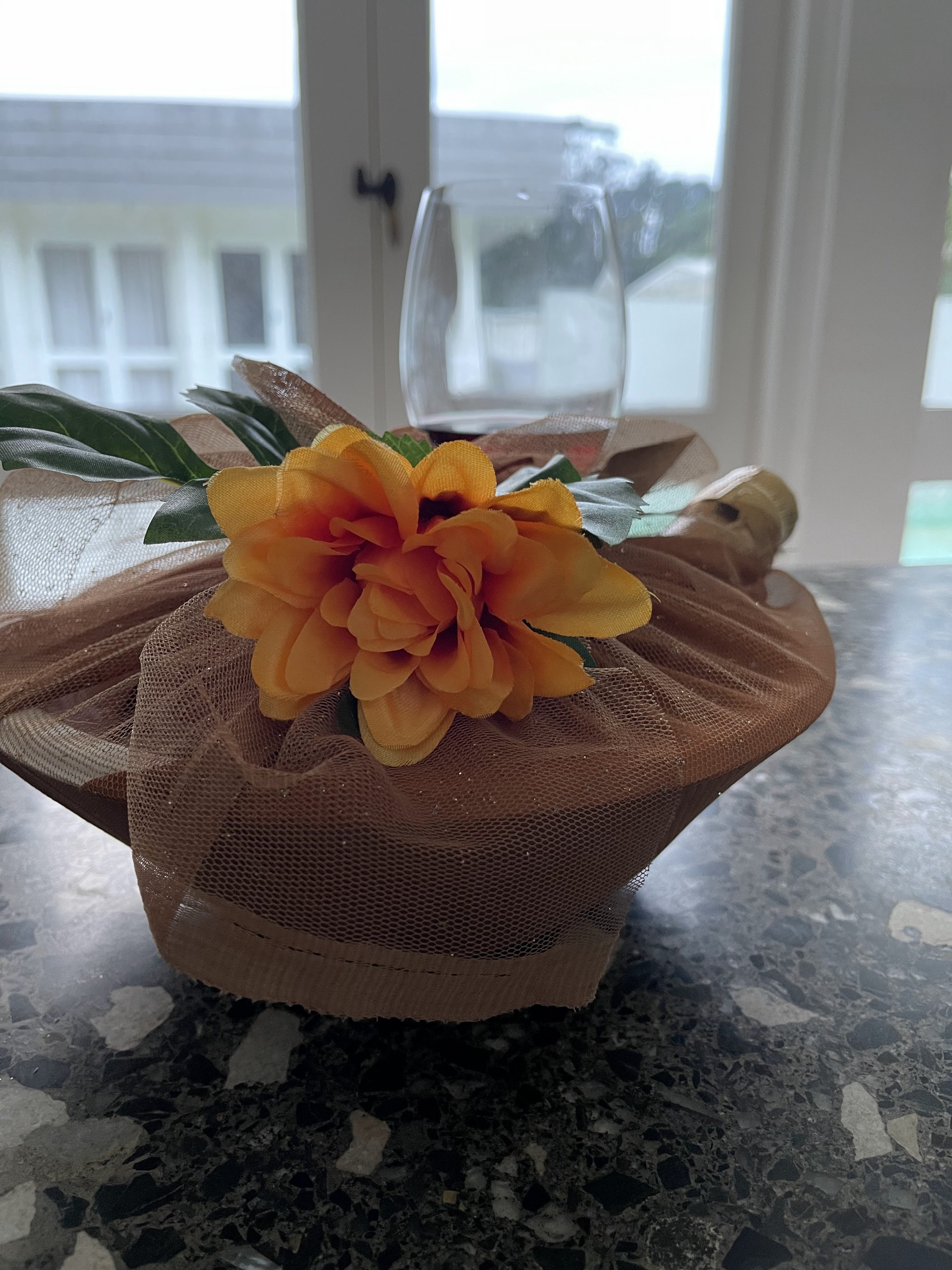 Yellow artificial flower with green leaves on a decorative basket with mesh wrapping, placed on a granite countertop with a blurred background of windows and a glass of wine.
