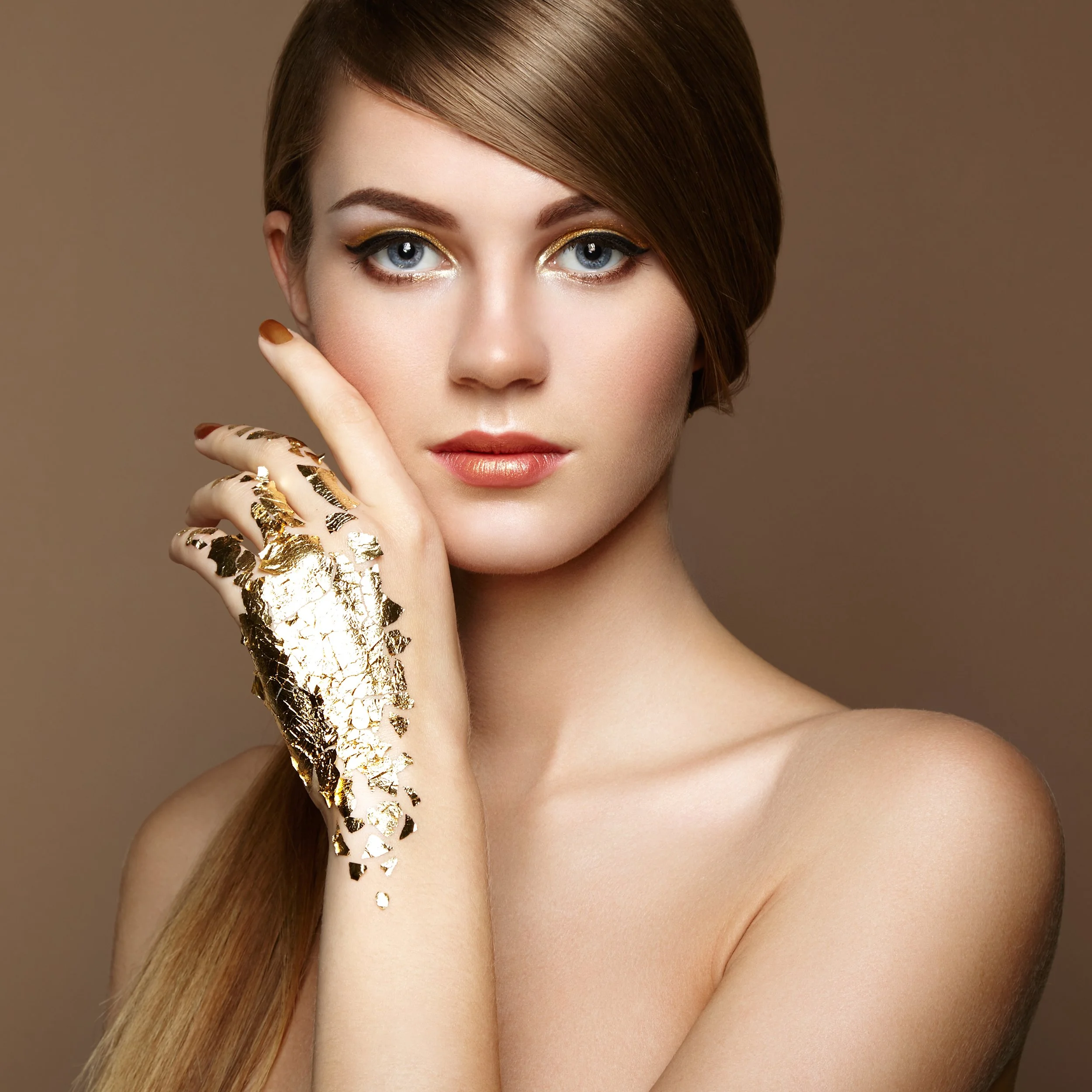 A woman with blue eyes has gold makeup on her eyelids and lips. She is touching her face which is covered with gold leaf, representing the anti-aging properties of gold, copper and silver minerals infused into Lira Clinical skincare products.