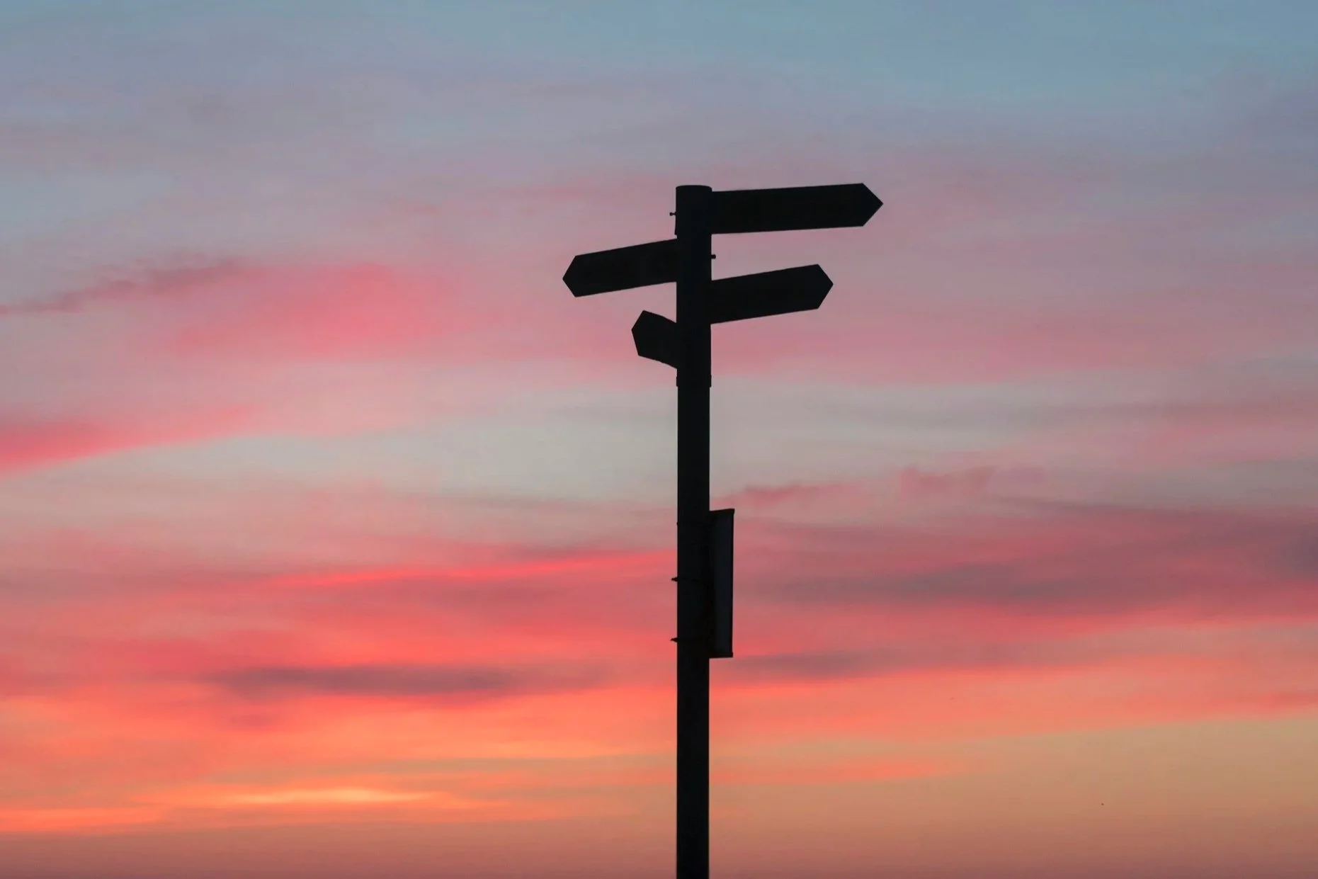 Un panneau de signalisation silhouetté contre un ciel coloré au coucher du soleil, avec plusieurs flèches pointant dans différentes directions.