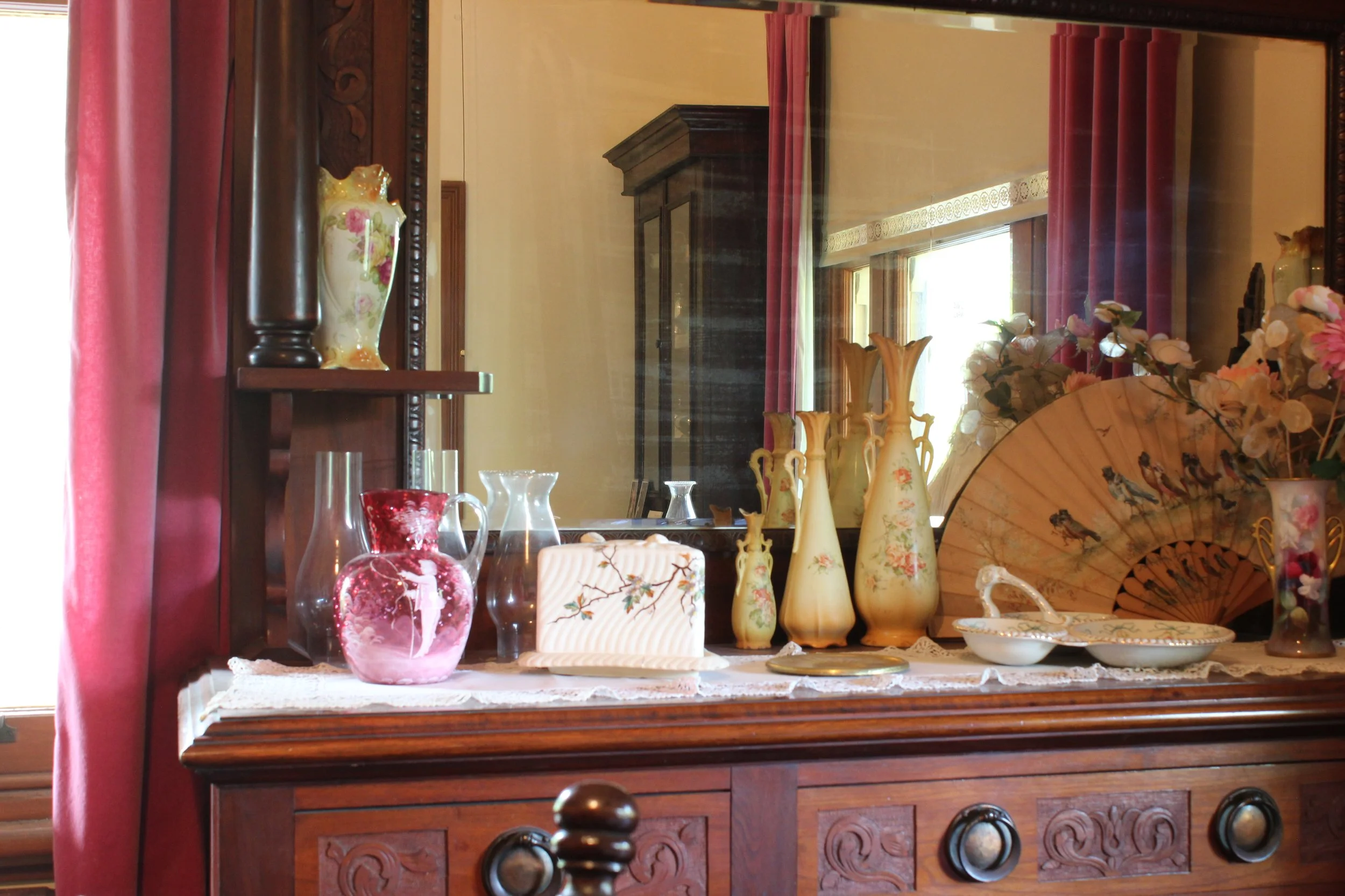 Decorative china and vases displayed on a wooden dresser in a vintage-style room with pink curtains and a large mirror.