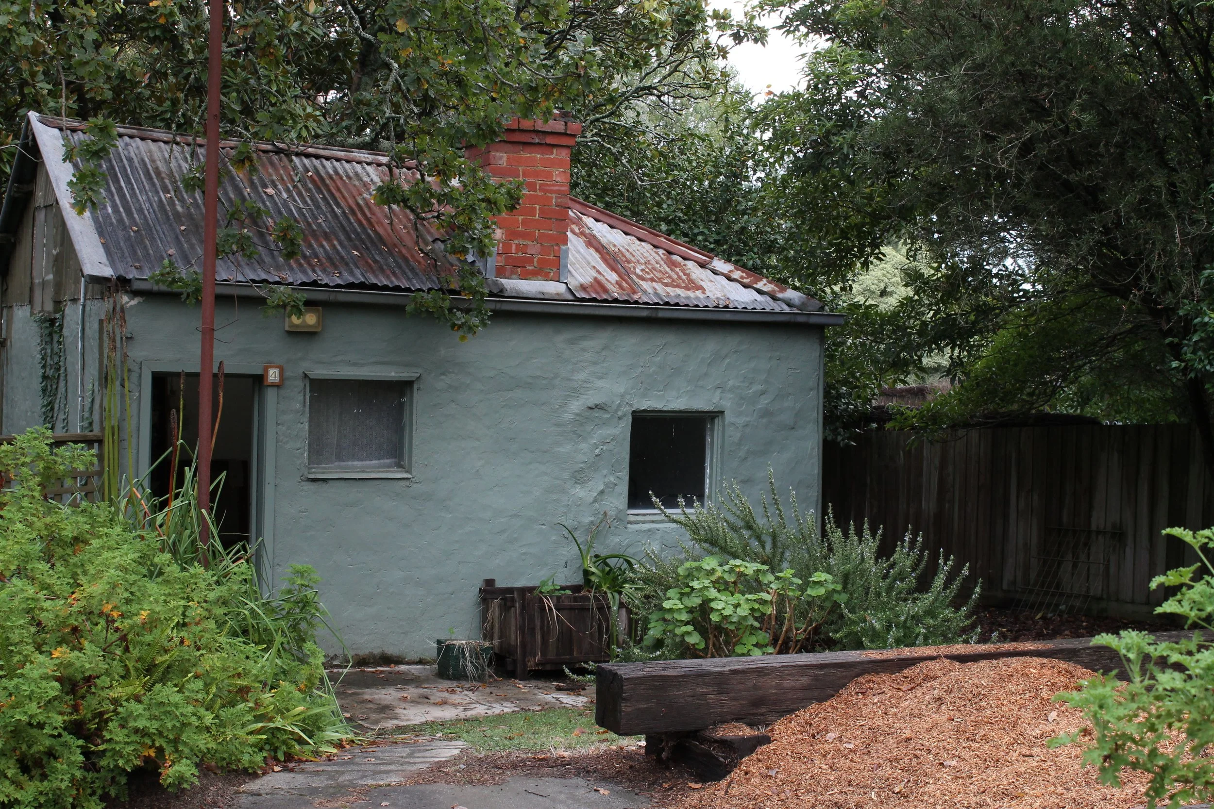 A small house with a green exterior, rusted metal roof, and a brick chimney. There are two windows and a door slightly ajar. The front yard has various plants, a wooden bench, and a mound of dirt or mulch. Trees and a wooden fence surround the house.
