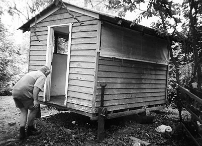 A person with a hat and shorts leans into a small wooden house on stilts, surrounded by trees in a forested area.