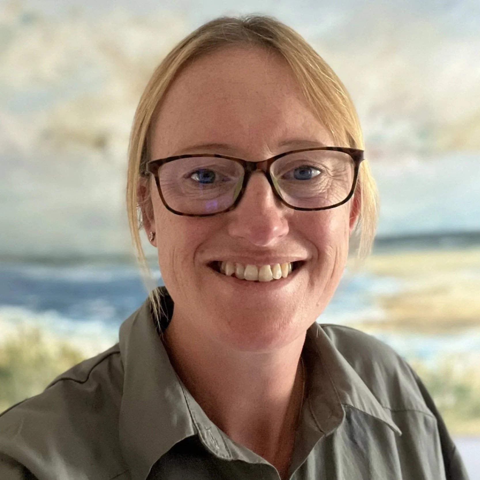 A smiling woman with glasses and short reddish hair, wearing a gray uniform, with a beach scene in the background.