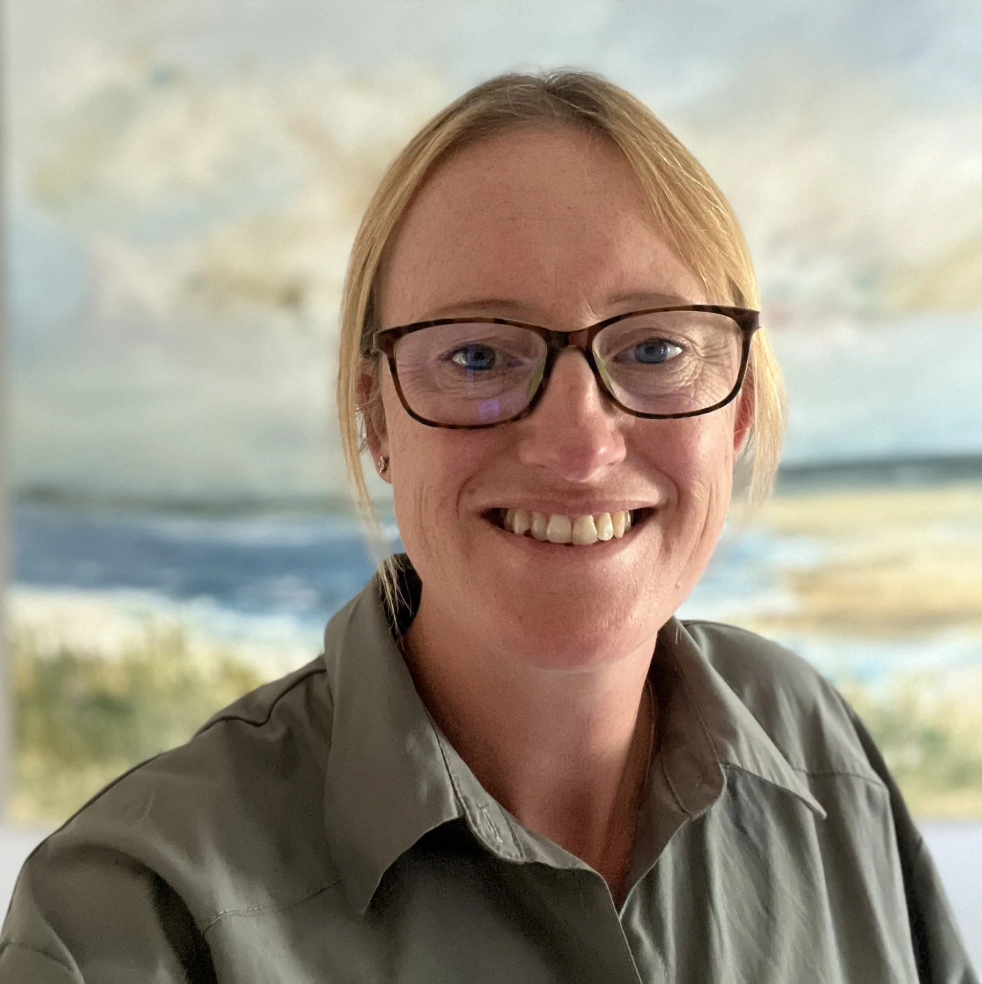 A smiling woman with glasses and short reddish hair, wearing a gray uniform, with a beach scene in the background.