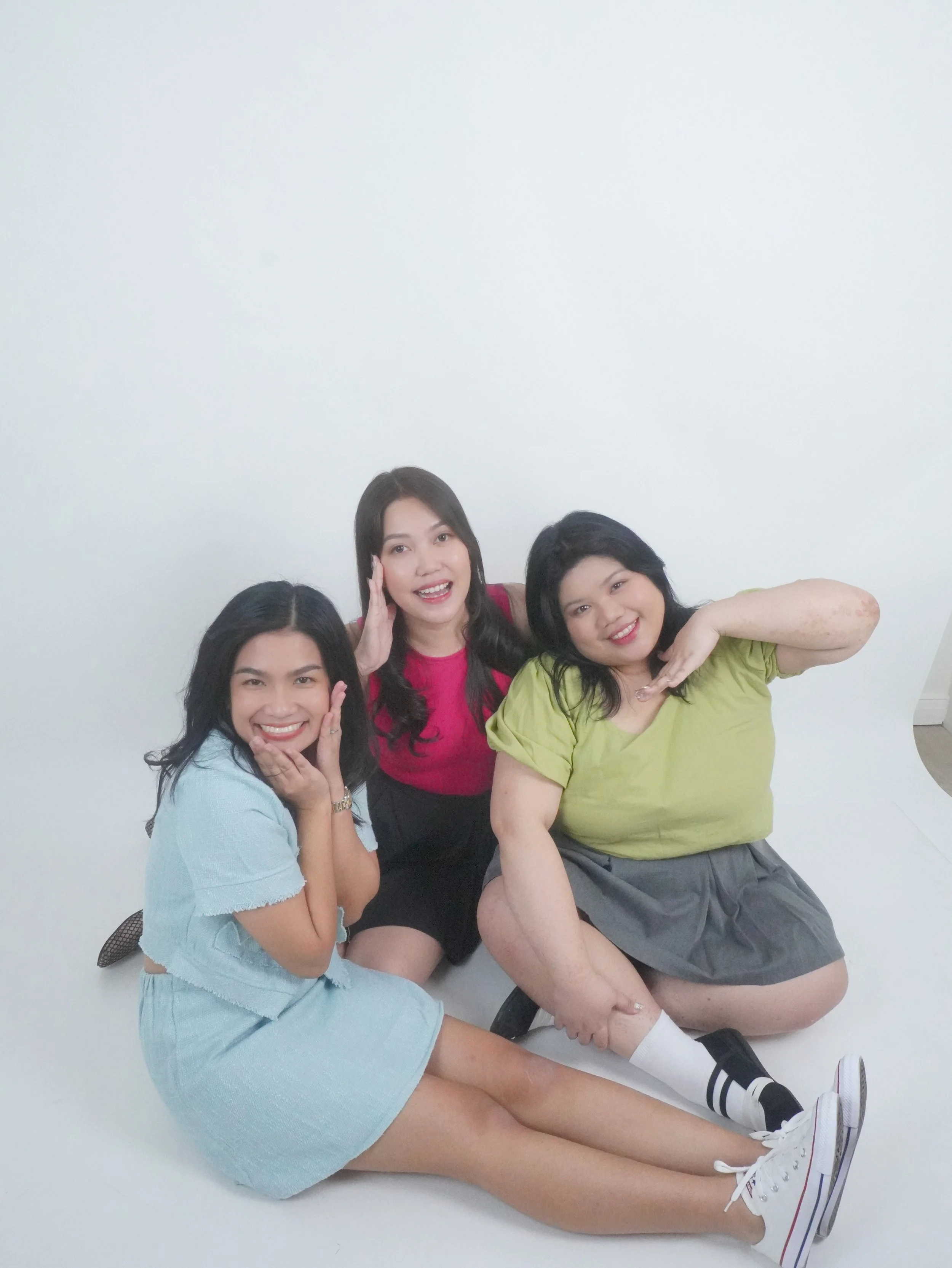 Three young women posing together on a white background, smiling and making playful gestures.