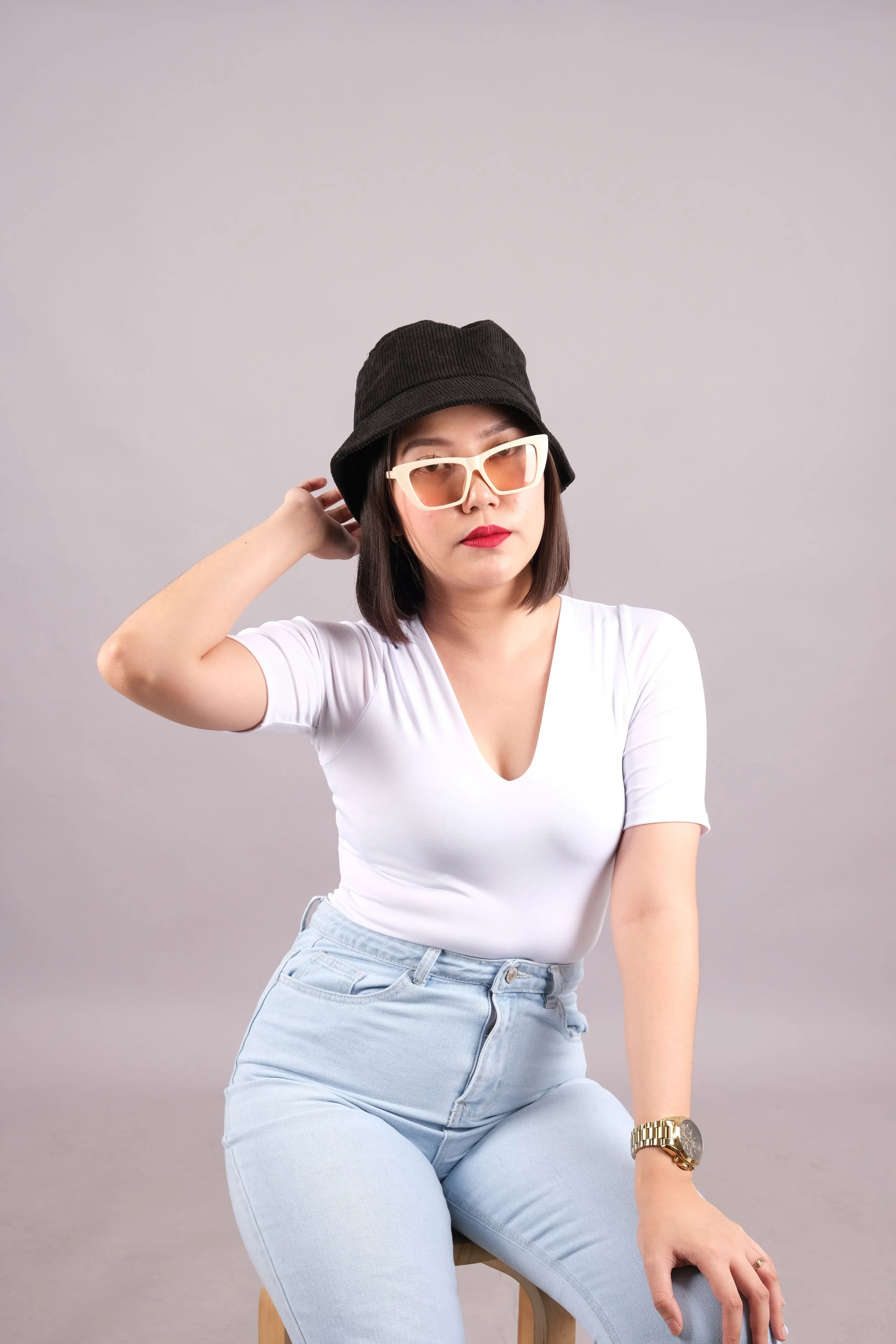A young woman with shoulder-length hair wearing a black bucket hat, white sunglasses, a white V-neck t-shirt, light blue jeans, a gold wristwatch, and red lipstick poses sitting on a stool against a plain gray background.