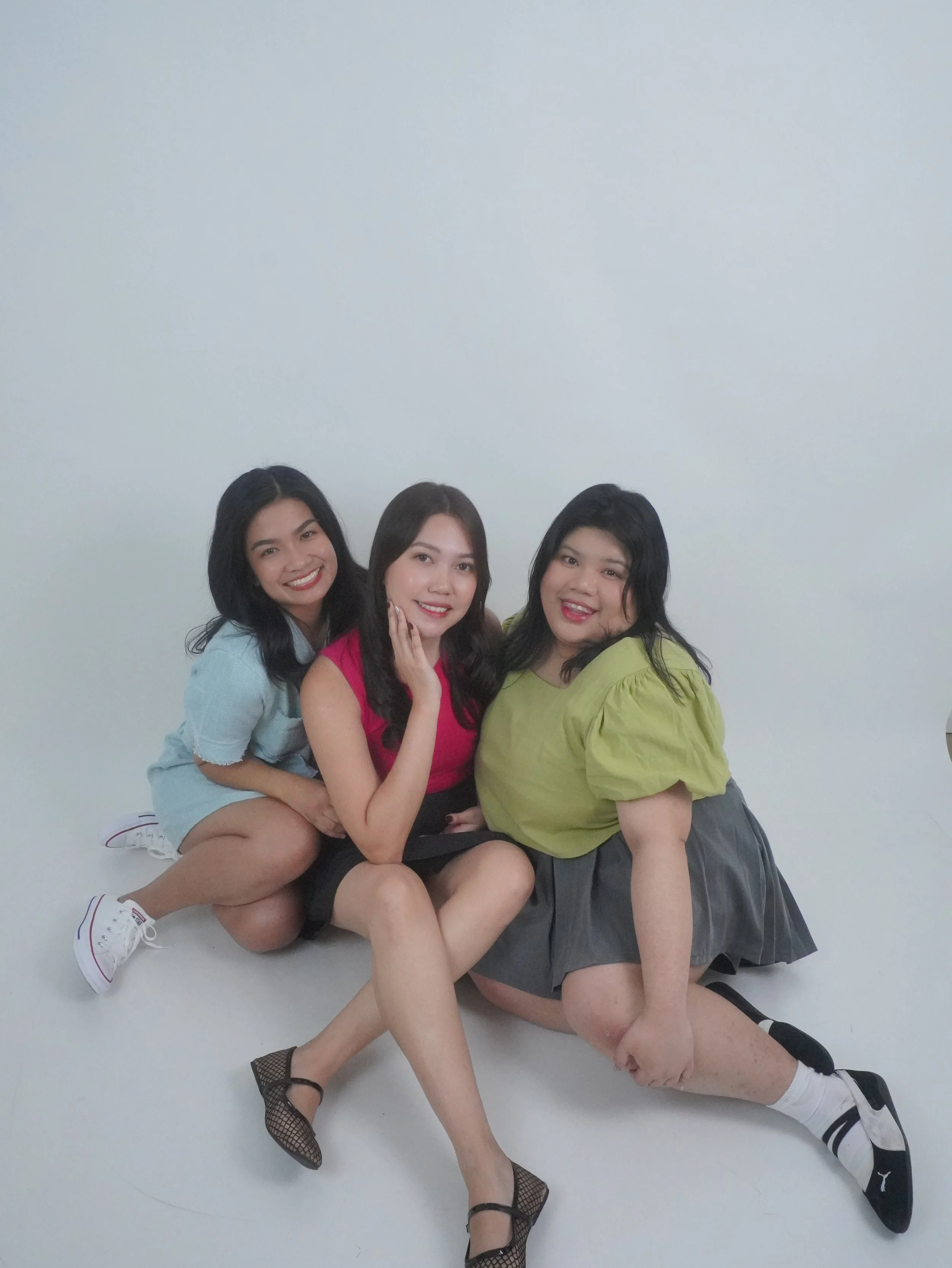 Three young women sitting on the floor together, smiling, posed in front of a plain white background. They are dressed casually with colorful tops and skirts or shorts, and wearing sneakers or shoes.