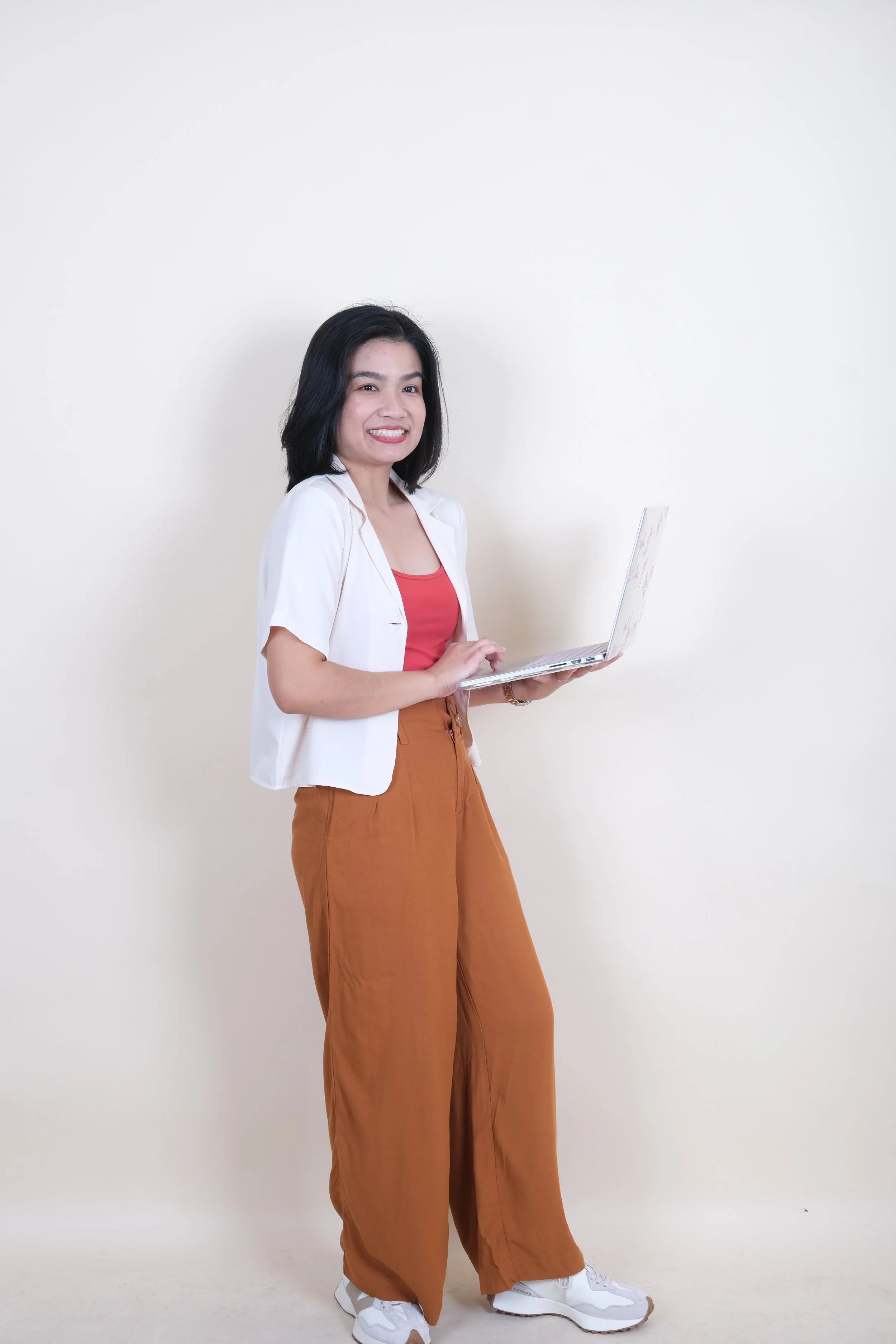 Woman standing against a plain white wall, holding a laptop, wearing a white short-sleeve shirt over a red top, beige wide-leg pants, and white sneakers, smiling at the camera.