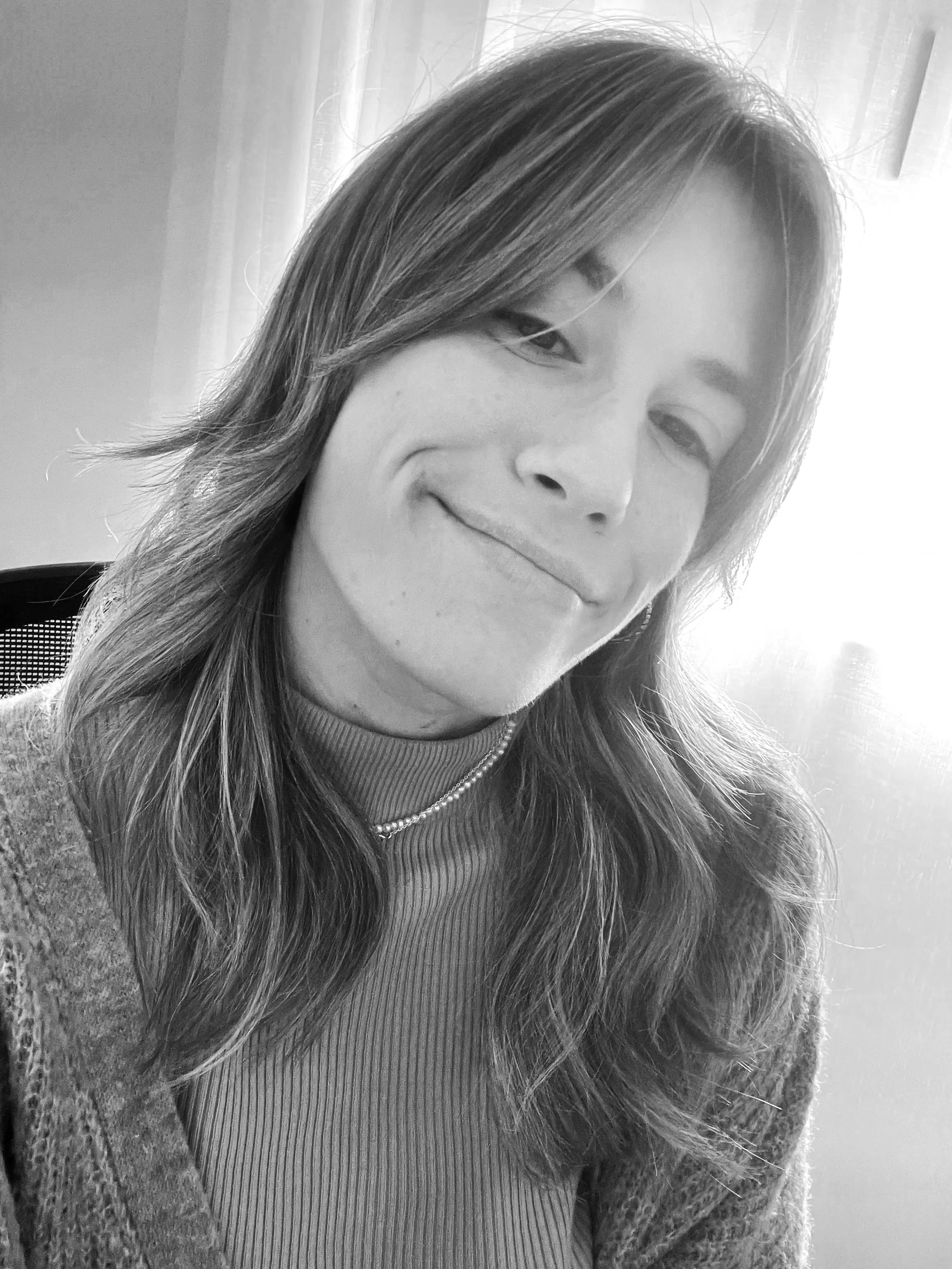 Black and white photo of a woman with shoulder-length hair, smiling slightly, sitting outdoors near a window with curtains.