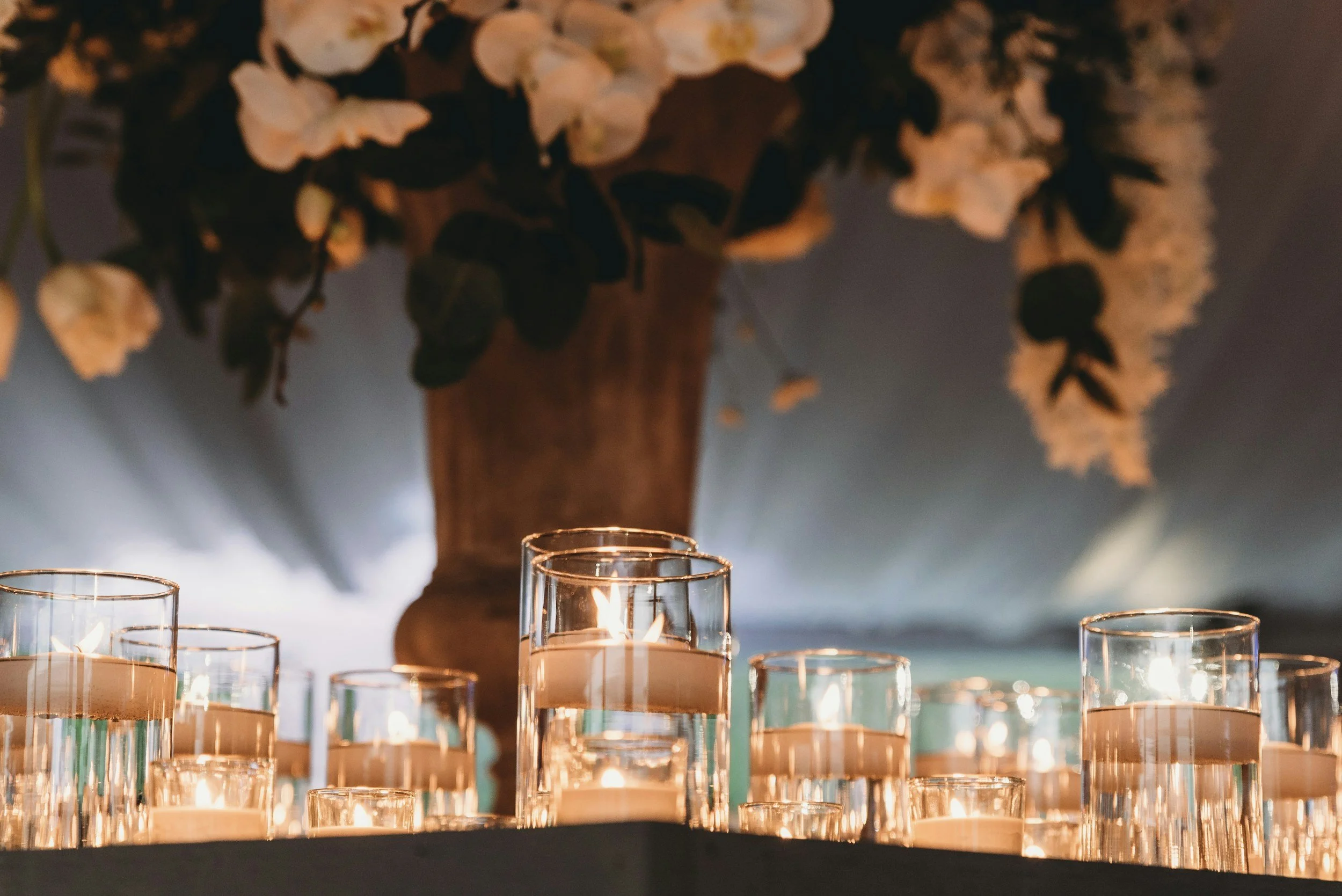 Mehrere Kerzen in Gläsern auf einem Tisch, mit einer großen Blumen- und Pflanzenarrangement in einem Vasen im Hintergrund, bei einer festlichen oder romantischen Gelegenheit.