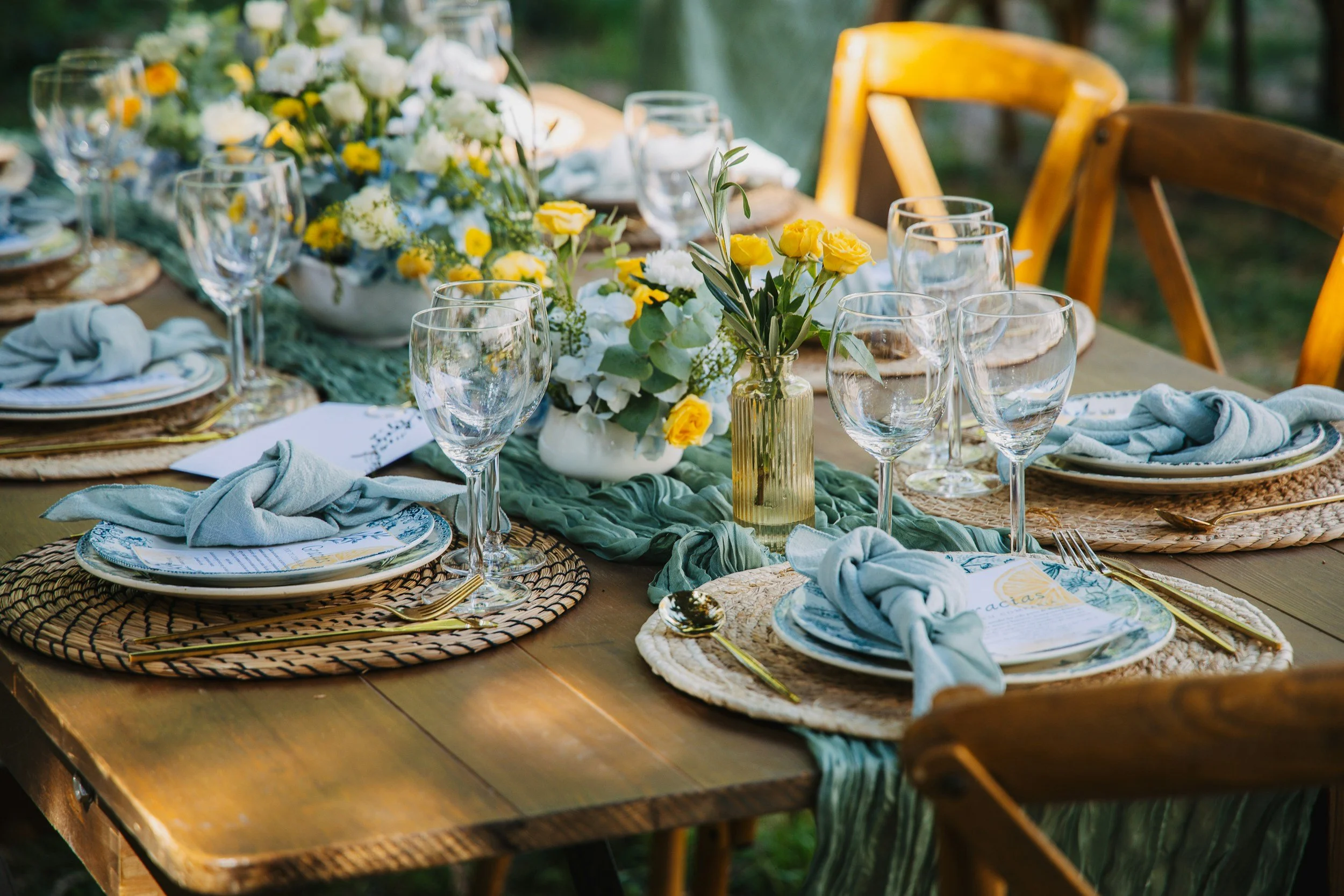 Ein festlich gedeckter Tisch mit blumiger Dekoration, Weingläsern, Tellern, Besteck und Stoffservietten, der für eine Outdoor-Feier vorbereitet ist.