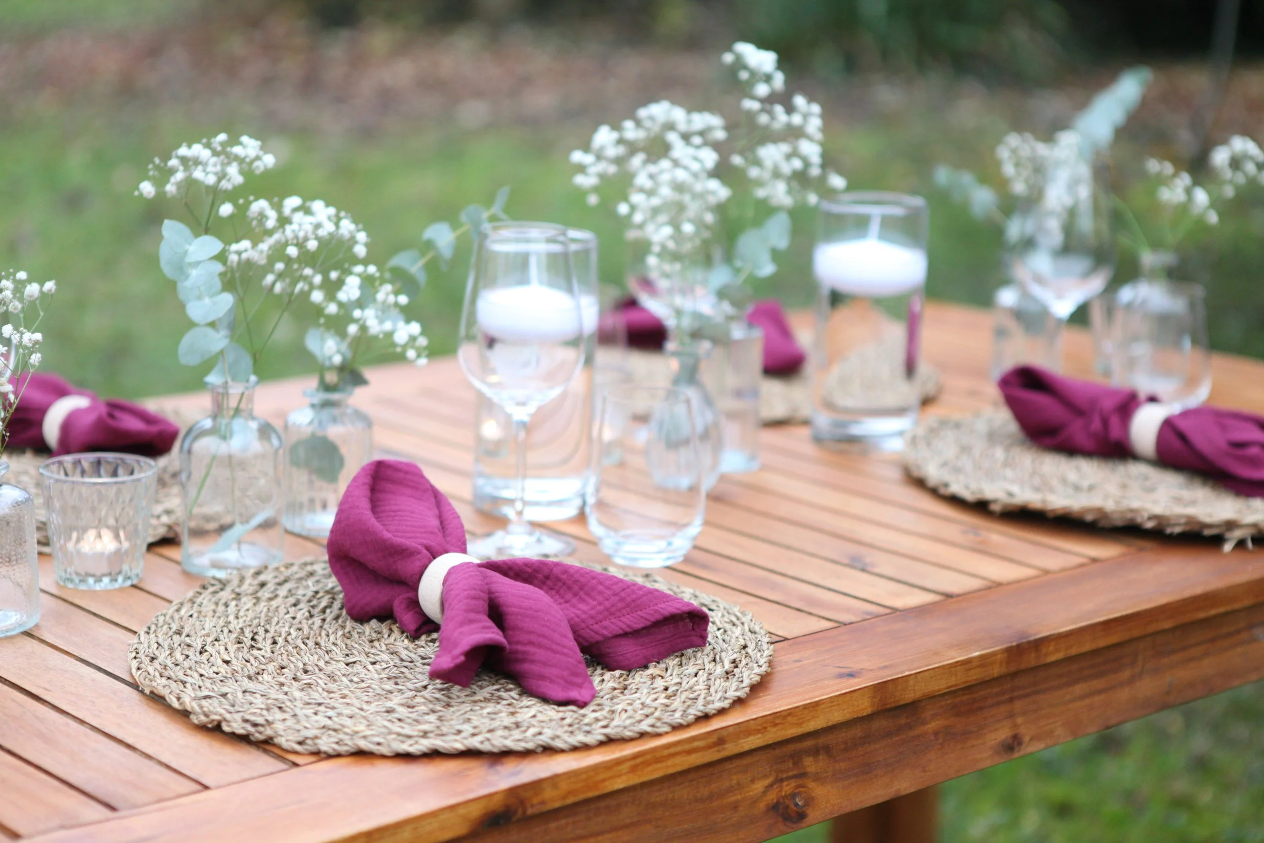 Ein gedeckter Holztisch im Garten mit Platzsets, Weingläsern, kleinen Vasen mit weißen Blumen und dunkelvioletten Stoffservietten