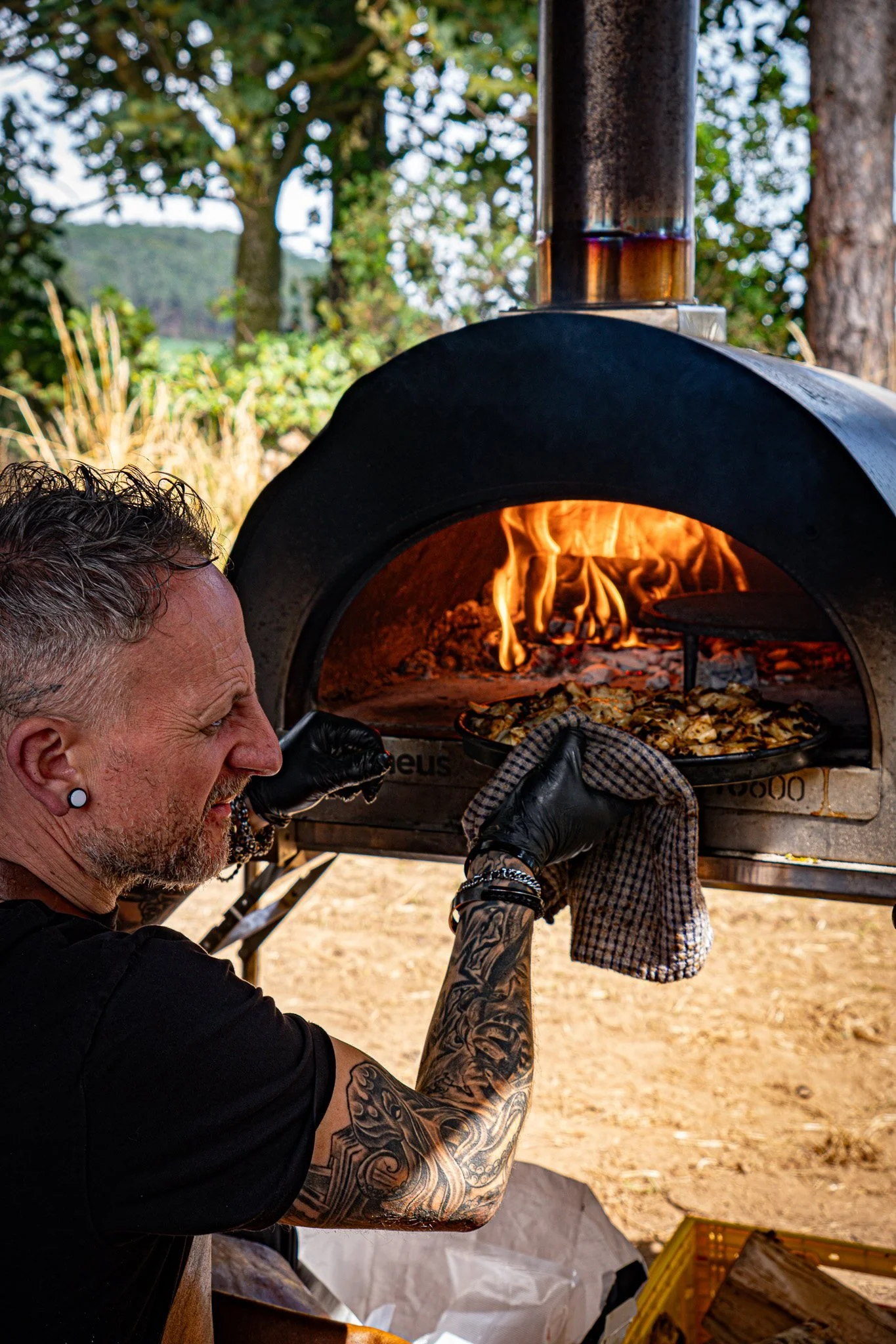 A man with tattoos and earrings standing outdoors, wearing black gloves, placing a pizza into a wood-fired oven with a flame inside.