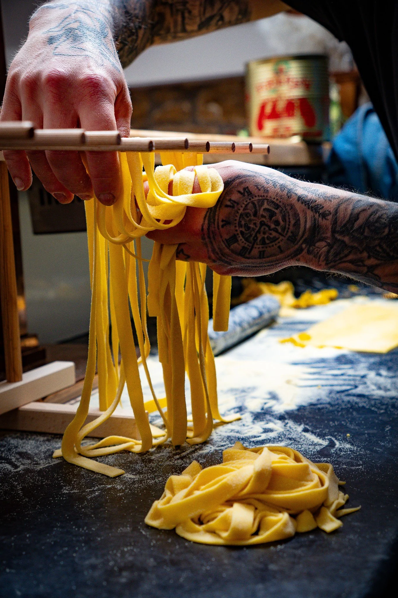 Person with tattoos on arms making fresh pasta, holding a fork to lift pasta strands over a dark surface with flour, with a wooden table and canned food in the background.